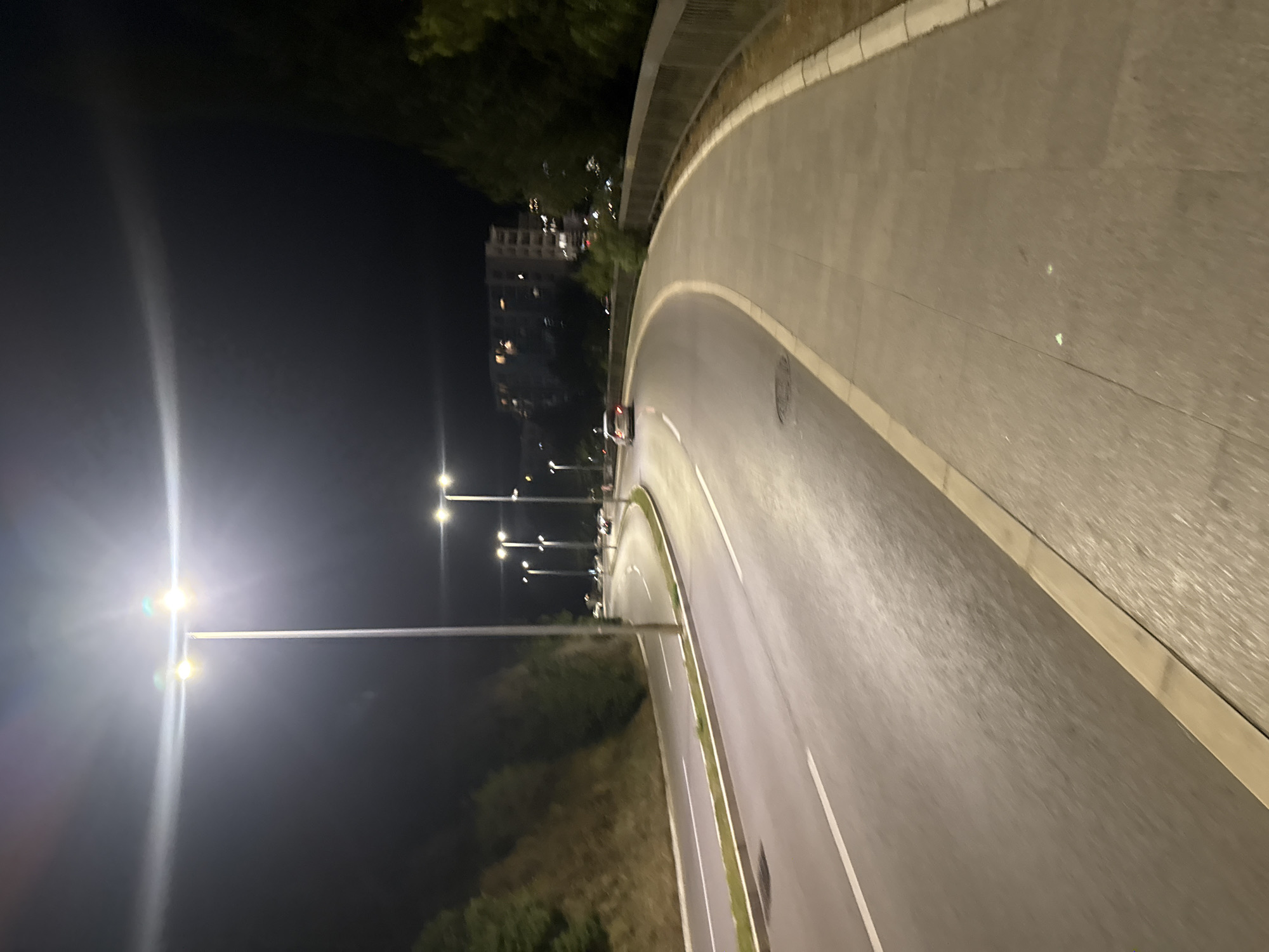This is a nighttime photograph of a curved road in the city of Boreti, Montenegro. The road is illuminated by streetlights, with several cars visible in the distance. A paved sidewalk runs alongside the road, bordered by a metal railing. Lush green trees and vegetation are present on the left side of the road, and buildings can be seen in the background. The overall atmosphere is calm and quiet, with the streetlights casting a warm glow on the asphalt.