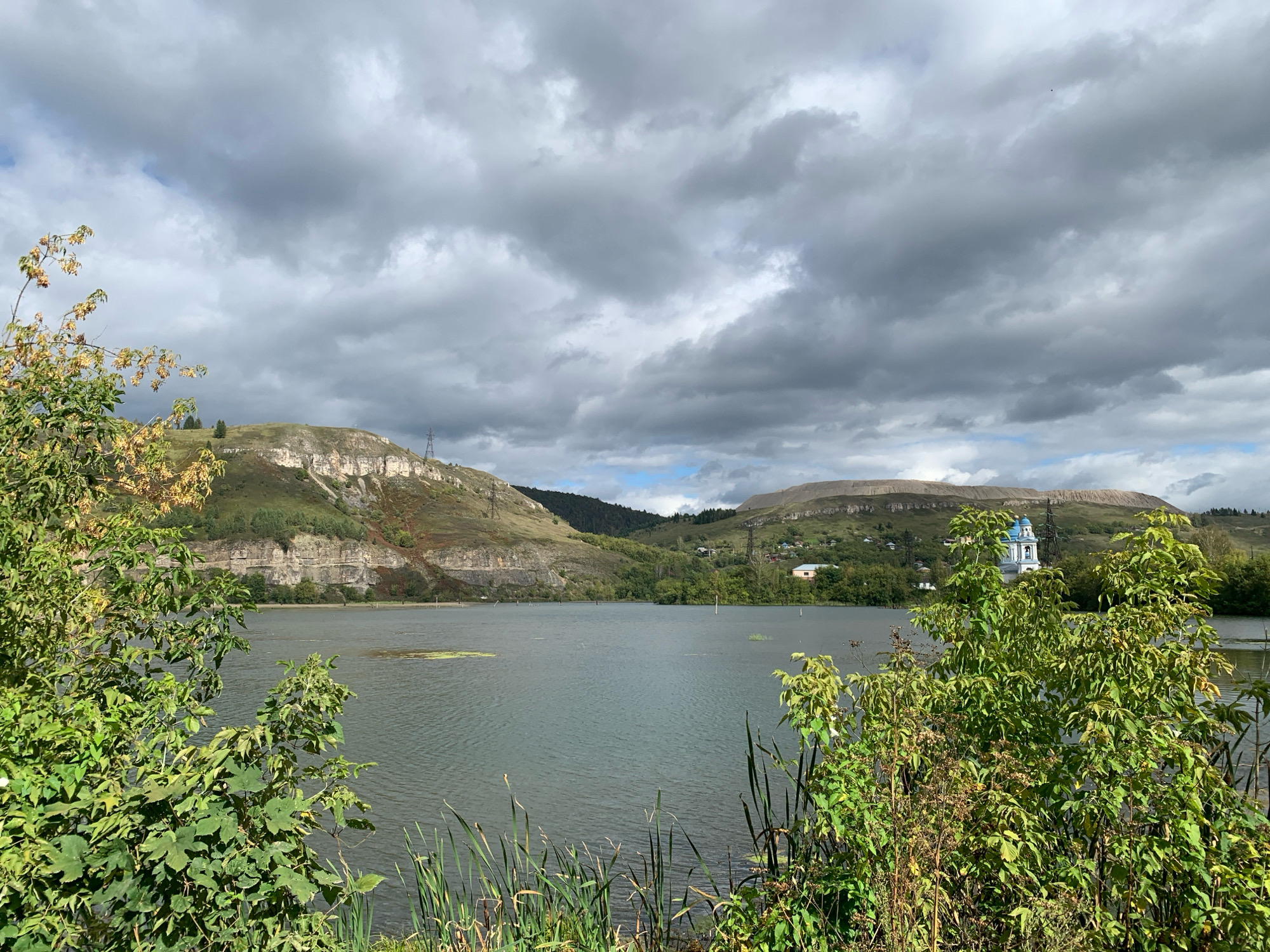 This is a medium-shot, landscape photograph taken on an overcast day in Minyar, Russia. The scene depicts a body of water, likely a river or lake, in the foreground and middle ground. The water is calm with slight ripples. Along the bank in the immediate foreground, tall green reeds and leafy green foliage are visible.
In the background, rolling hills covered in greenery rise from the far bank of the water. The hills are uneven, with some sections showing exposed rock faces. Several small buildings, including a blue-domed church, are nestled among the trees on the slopes. Power lines and towers are also present on the hills. The sky is filled with heavy, dark grey clouds, with hints of blue sky peeking through in some areas. The overall impression is a natural landscape with evidence of human habitation.