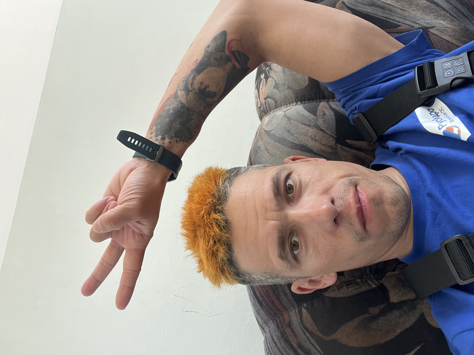 A man with dyed orange hair is lying on a patterned sofa. He is wearing a blue shirt with the words "RYCHARDO Bomercay3C" on it, and a black strap is visible across his chest. His left arm is raised, displaying a tattoo of a dog. He is wearing a black smartwatch. The background consists of a plain white wall. The photo appears to be taken in the city of Chaikovsky, Russia, based on the Cyrillic script on the shirt.