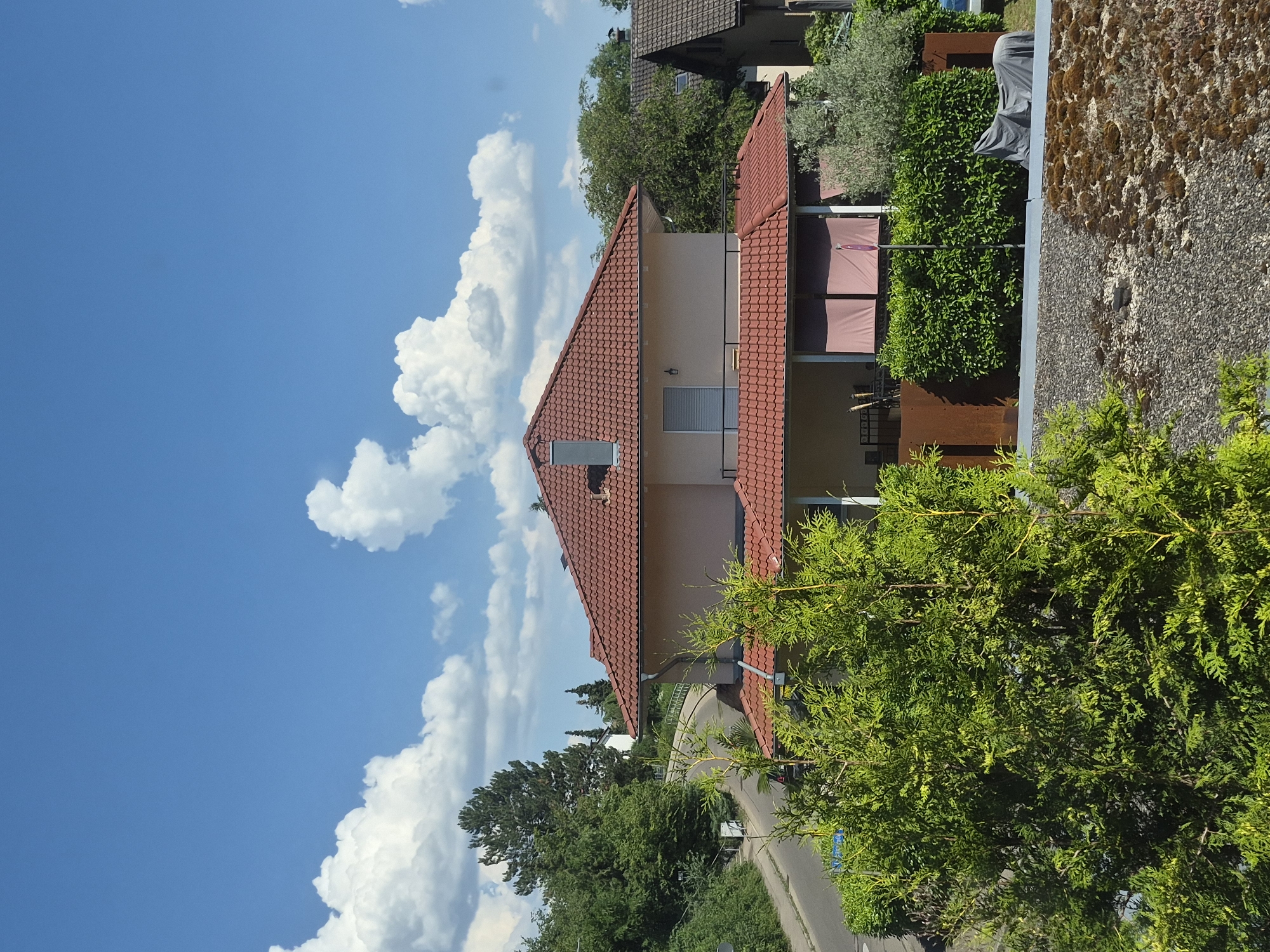 This is a vertical, eye-level, full shot of a residential area under a bright blue sky with fluffy white clouds. The scene is dominated by a house with a red tiled roof and light-colored walls. A chimney protrudes from the roof. In front of the house, there is a covered porch area with what appear to be retractable awnings. The house is surrounded by lush green trees and bushes. To the right of the house, a weathered wall made of stones and gravel is visible. A portion of a road with a light-colored surface runs in front of the house and disappears into the greenery. The image appears to have been taken on a sunny day, suggesting fair weather. The overall mood is peaceful and tranquil, typical of a suburban environment. There is no visible text in the image.