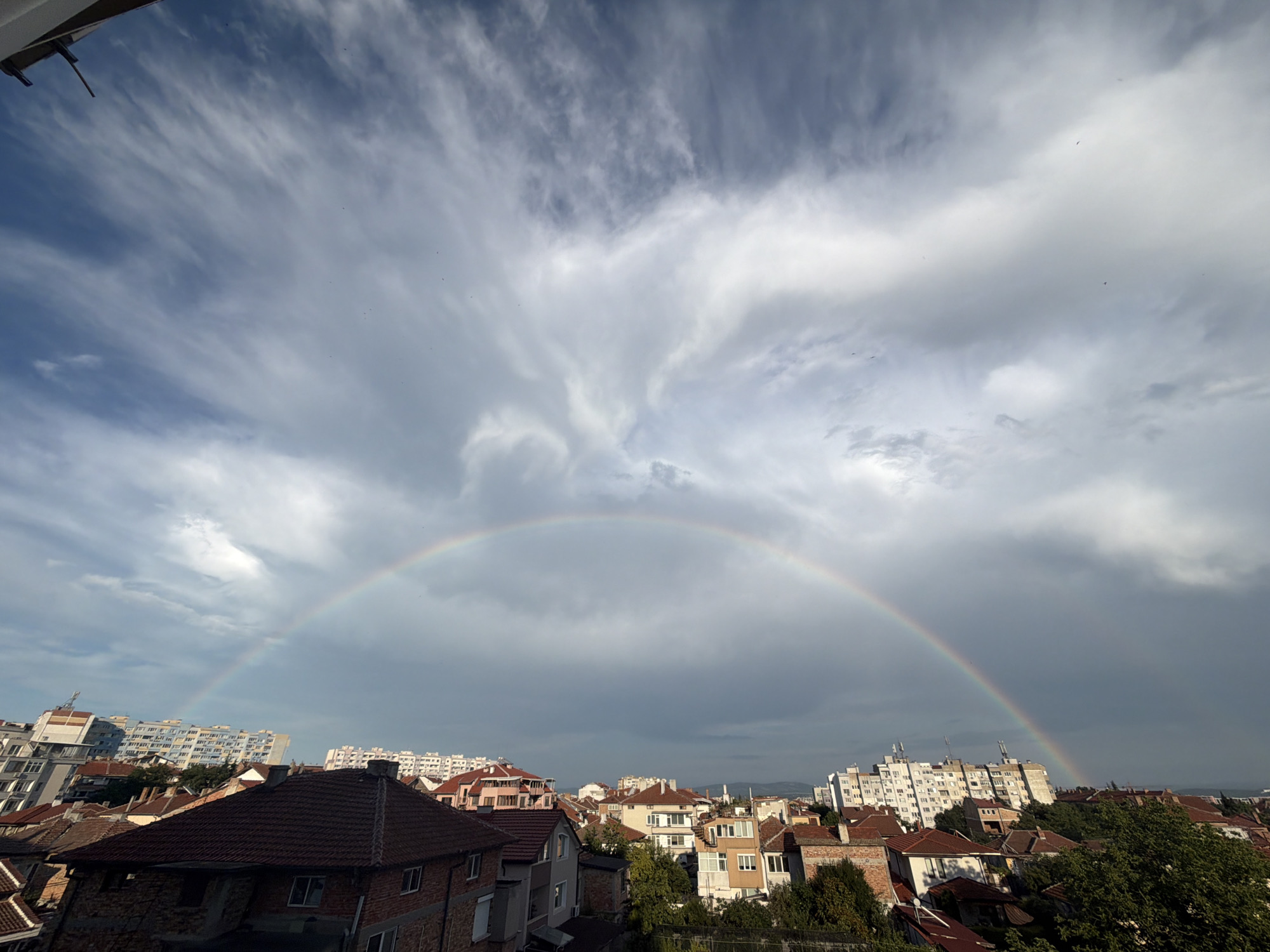 The image depicts a cityscape in Burgas, Bulgaria, under a dramatic sky featuring a prominent rainbow. The perspective is from an elevated vantage point, looking out over a dense collection of buildings. In the foreground, the tiled roofs of residential houses are visible, some with exposed brickwork. Further back, multi-story apartment buildings dominate the skyline, characterized by their varied architectural styles and light-colored facades. Trees and greenery are interspersed among the buildings, adding a touch of nature to the urban environment. The sky is a dynamic mix of blue and grey clouds, with streaks of cirrus clouds high above, creating a sense of depth and movement. The rainbow arches across the frame, its vibrant colors contrasting with the subdued tones of the clouds and city. There are no visible people or animals in the scene. The overall impression is one of a post-rainy day, with the natural phenomenon of a rainbow offering a striking visual element against the backdrop of urban life.