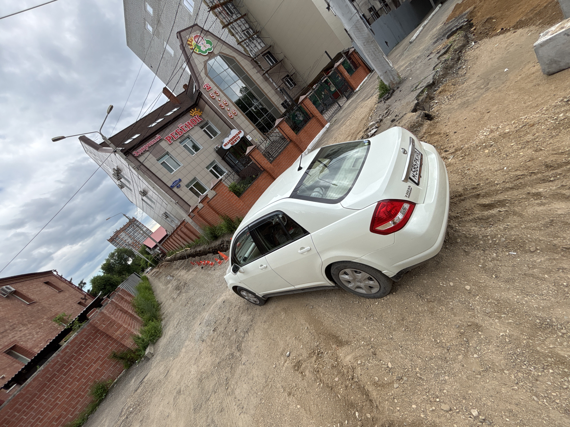 A white Nissan Tiida sedan is parked on a dirt and gravel road in front of a multi-story building in Blagoveshchensk, Russia. The road appears to be under construction or in disrepair, with a section of pavement eroded away on the right side of the image. The building has a red brick facade and is adorned with signage in Russian, including "Здоровый ребёнок" which translates to "Healthy Child". Orange traffic cones are visible along the edge of the eroded road. The sky is overcast with clouds.