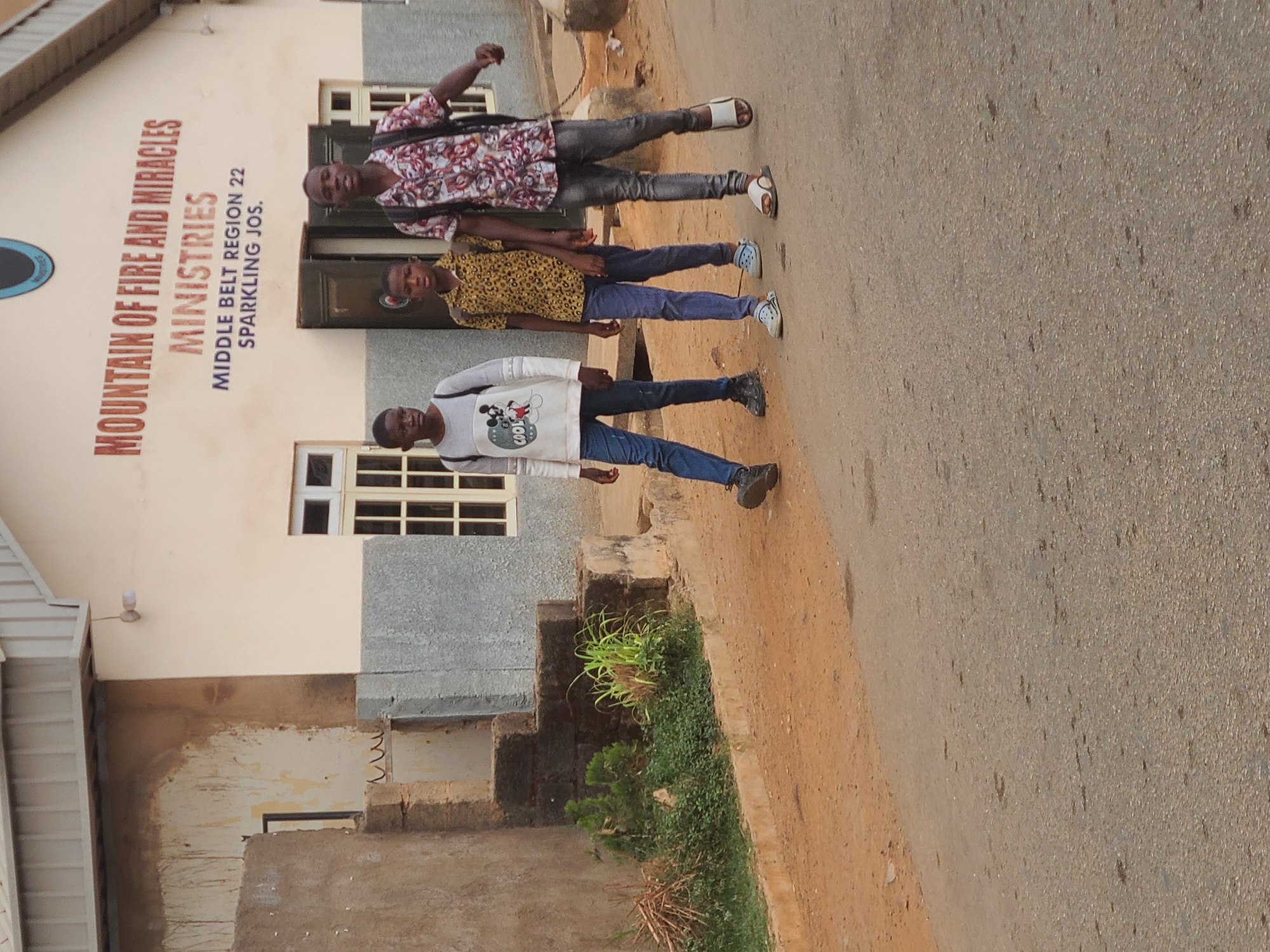 Three young men stand on the edge of a paved road next to a beige and grey building. The building is the Mountain of Fire and Miracles Ministries, Middle Belt Region 22 Sparkling Jos, located in the city of Jos, Nigeria. The men are arranged in a line, with the first wearing a white Mickey Mouse sweater and blue jeans, the second wearing a yellow leopard-print shirt and blue jeans, and the third wearing a patterned shirt and grey jeans. They are all facing the camera. The weather is bright and sunny.