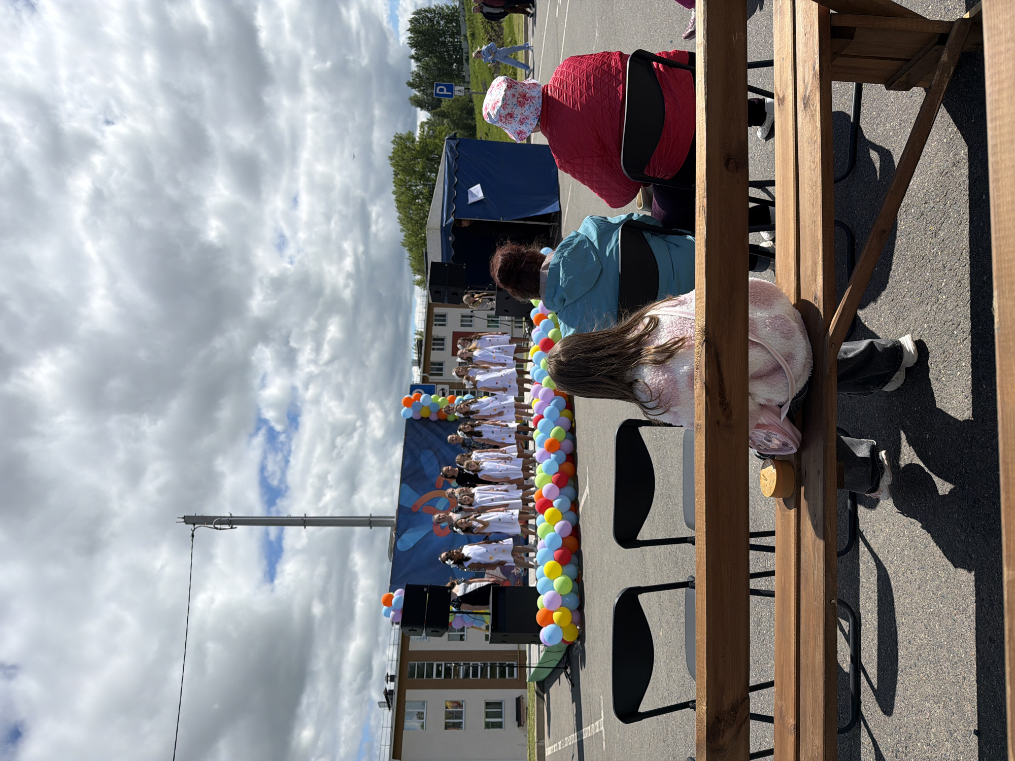 The image, taken in the city of Valozhyn, Belarus, depicts an outdoor event. A group of young people, presumably dancers, are performing on a stage decorated with balloons and a backdrop featuring a graphic image. The stage has speakers positioned in front. Several people are watching the performance, seated on chairs. The scene is partially viewed from behind a wooden structure. The weather is overcast. A building is visible in the background.