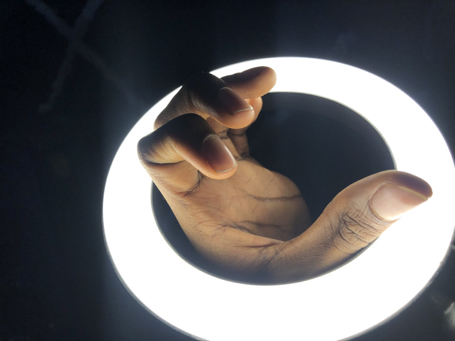 The image displays a human hand with a dark skin tone, centrally positioned within an illuminated circular ring light. The hand is partially open, with fingers slightly flexed and the palm oriented generally forward. Visible details include fingernails and natural creases on the skin. The ring light emits a bright white, uniform illumination, providing the sole light source against an otherwise dark and featureless background. No other individuals, objects, or environmental elements are discernible. This scene is situated in Tema, Ghana.