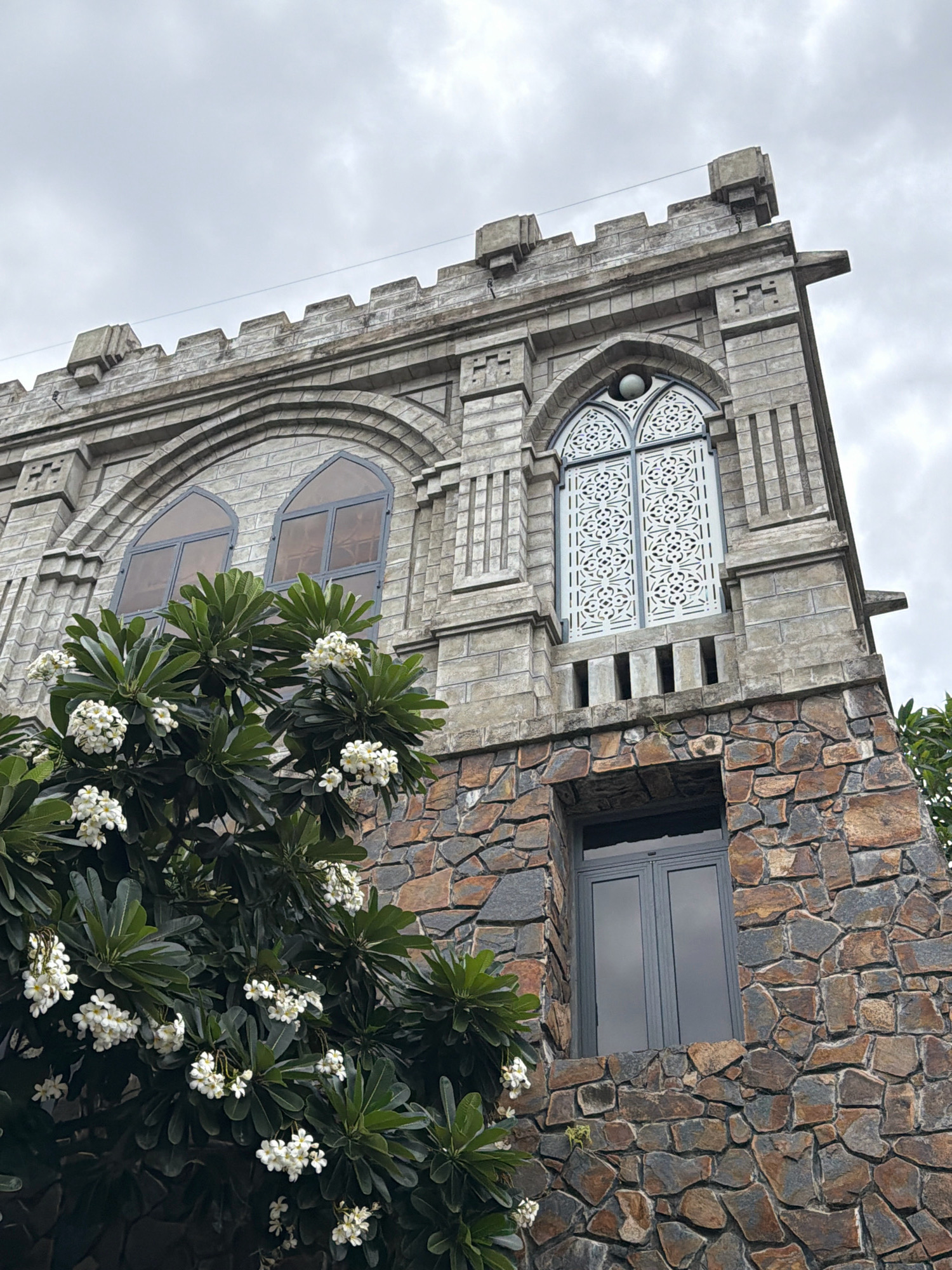 This is a low-angle shot of the exterior of a building in Nha Trang, Vietnam, with a cloudy sky overhead. The building's architecture features a stone base and a facade constructed of grey blocks, with battlements along the roofline. There are arched windows with decorative white grilles and a large, dark-paned window on the lower stone level. In the foreground, a lush green tree with clusters of white flowers, likely frangipani, partially obscures the building. The image is devoid of people or any discernible activity.