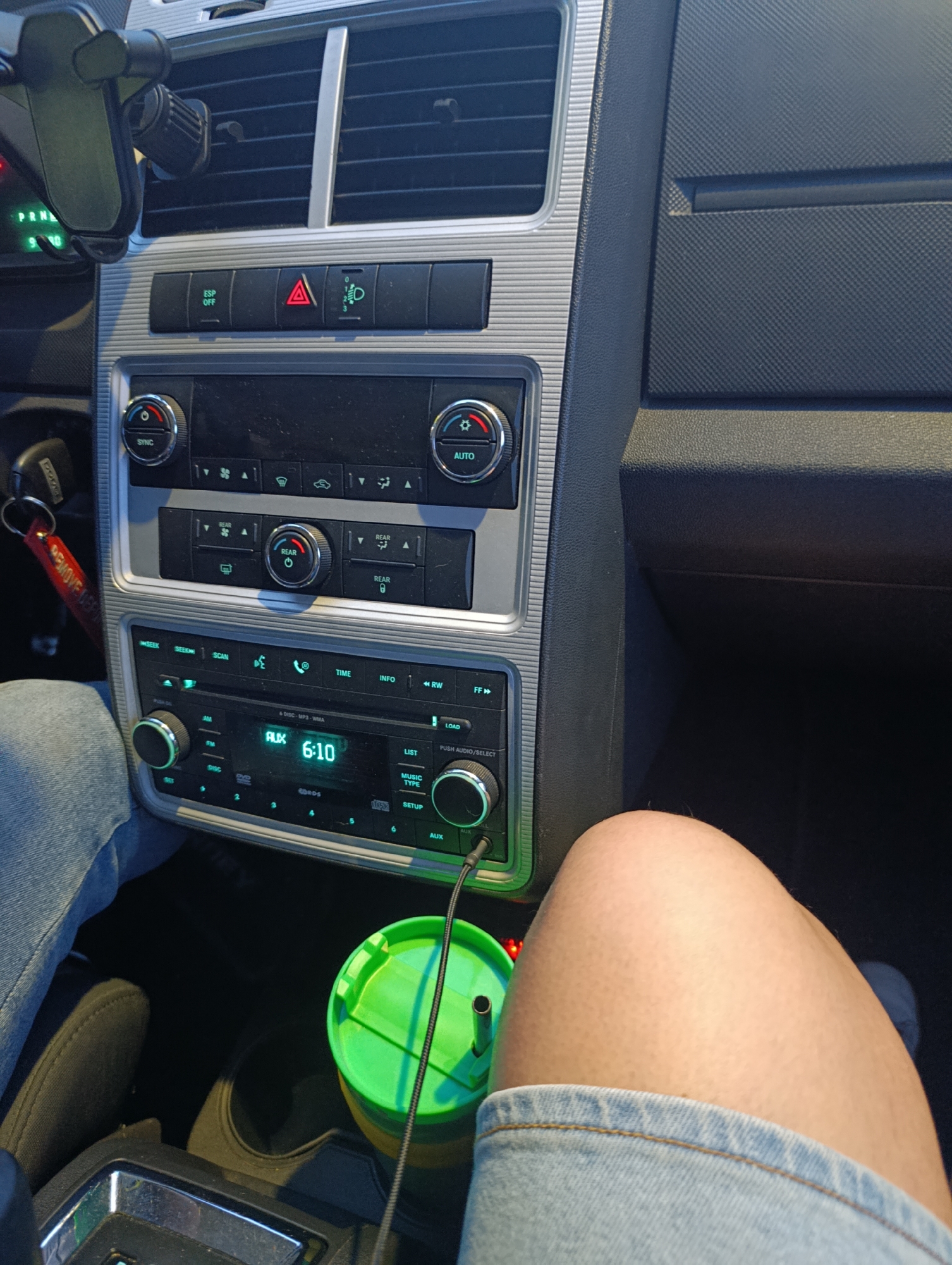 The image captures the interior of a car, focusing on the center console, dashboard, and parts of a person's legs. The setting appears to be during daytime, with the car likely stationary or parked, as indicated by the illuminated "PRND" text (suggesting Park or Neutral) near the gear shifter.
In the foreground, the upper legs of a person are visible, wearing light-wash denim shorts or jeans. A bright green tumbler with a lid and straw is placed in a cup holder, connected by a black audio cable to the car's AUX port.
The car's dashboard and center console feature a silver-trimmed design. Air vents are visible at the top. Below them, buttons include "ESP OFF", a red hazard light triangle, and a headlight adjustment symbol. The climate control system comprises "SYNC" and "AUTO" dials, along with buttons for fan speed, air direction, and "REAR" controls.
The car's integrated radio and CD/DVD player is prominent, displaying "AUX" mode and the time "6:10". Its control panel includes buttons such as "AM", "FM", "DISC", "SEEK", "SCAN", "TIME", "INFO", "RW", "FF", "LIST", "MUSIC TYPE", "SETUP", and "LOAD". The screen also indicates "& DISC-MP3-WMA" capability. A car key, attached to a distinctive red "REMOVE BEFORE FLIGHT" keychain, hangs near the ignition. A phone mount is visible near the air vent on the left.
The scene depicts a typical car interior in Funza, Colombia, suggesting the person might be a passenger or driver waiting, with audio playing via the AUX input.