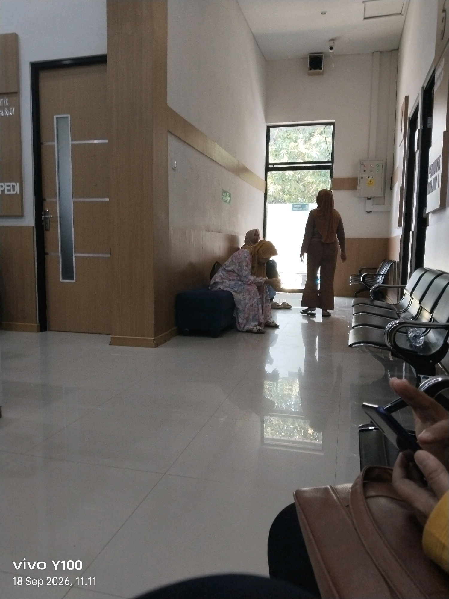 The image depicts a waiting area, possibly in a medical facility, with a tiled floor and beige walls. A large wooden door is on the left, with a vertical glass panel. A sign on the wall to the left of the door reads "PEDI." In the background, a glass door leads outside to a green, leafy environment.
There are three individuals visible in the waiting area. Two women are seated on a dark blue bench on the left side of the hallway. One is wearing a floral-patterned dress and a yellow headscarf, and the other, partially obscured, wears a dark garment and a beige headscarf. A third woman, wearing a beige hijab and a matching outfit, stands near the glass door, facing away from the camera.
To the right of the hallway, several rows of black and silver chairs are arranged. A person's hand holding a dark-colored mobile phone is visible in the foreground on the right. Above the chairs, a security camera is mounted on the wall. Electrical outlets and a control panel are also visible on the wall.
The image is dated "18 Sep 2026, 11.11" and includes a "vivo Y100" watermark, indicating it was captured with a Vivo Y100 smartphone.