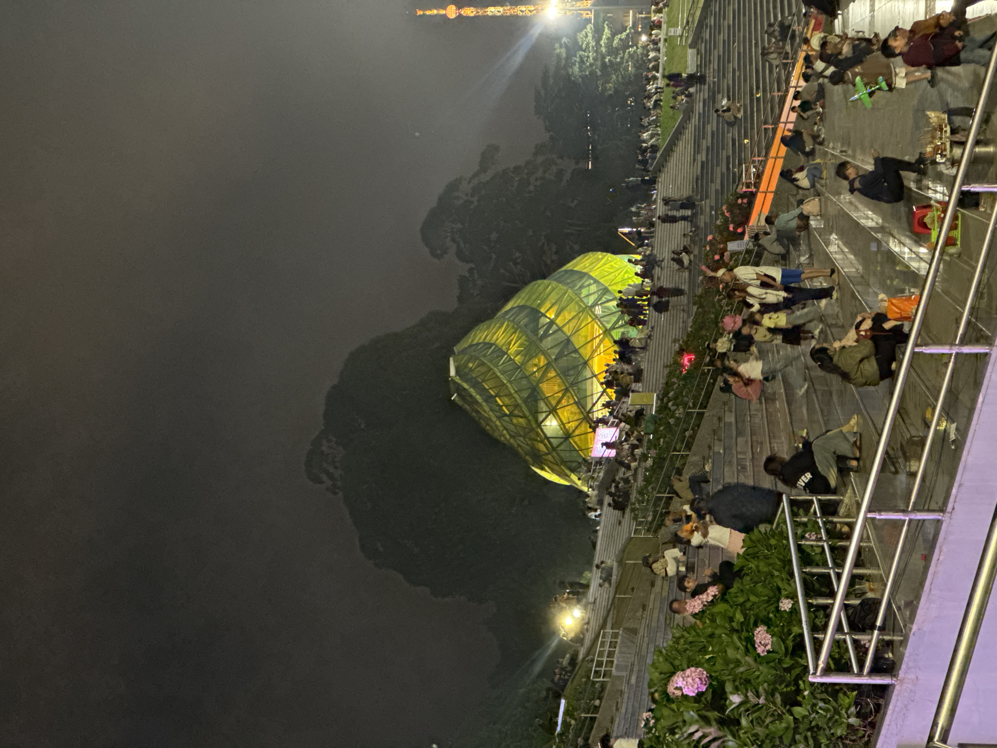 The image captures a vibrant night scene at an outdoor public area, likely a plaza or amphitheater, with a distinctive modern building and numerous people. The primary subjects include a large crowd of individuals, a prominent architectural structure, and various elements of the urban landscape.
The setting is illuminated by artificial lights, indicating it is nighttime. The sky is dark and a bit hazy, with distant lights piercing through, including what appears to be a brightly lit tower or structure in the upper background.
The most striking object is a large, glass-paneled building with a curved, geometric design, glowing with a bright yellow-green light. Its unique shape suggests a significant landmark, resembling the "Big Artichoke" building at Lam Vien Square in Da Lat, Vietnam, aligning with the provided location context.
People are gathered extensively on tiered concrete steps or bleachers that rise from the foreground towards the illuminated structure. They are seen sitting, standing, and reclining in various casual postures, suggesting a relaxed social atmosphere. Some individuals are facing the building, while others are interacting among themselves. The activity appears to be leisure-oriented, with people enjoying the evening ambiance, possibly watching a performance or simply congregating in the lively space. Green foliage and flower beds, featuring pink hydrangeas, are interspersed along the steps, adding natural elements to the urban environment.
Notable details include reflections of lights on surfaces, the sheer number of people creating a bustling crowd, and the distinctive architecture. On the back of one person sitting on the steps, the word "LOVER" is visible on their clothing. The overall mood appears pleasant and social, indicative of a popular evening gathering spot.