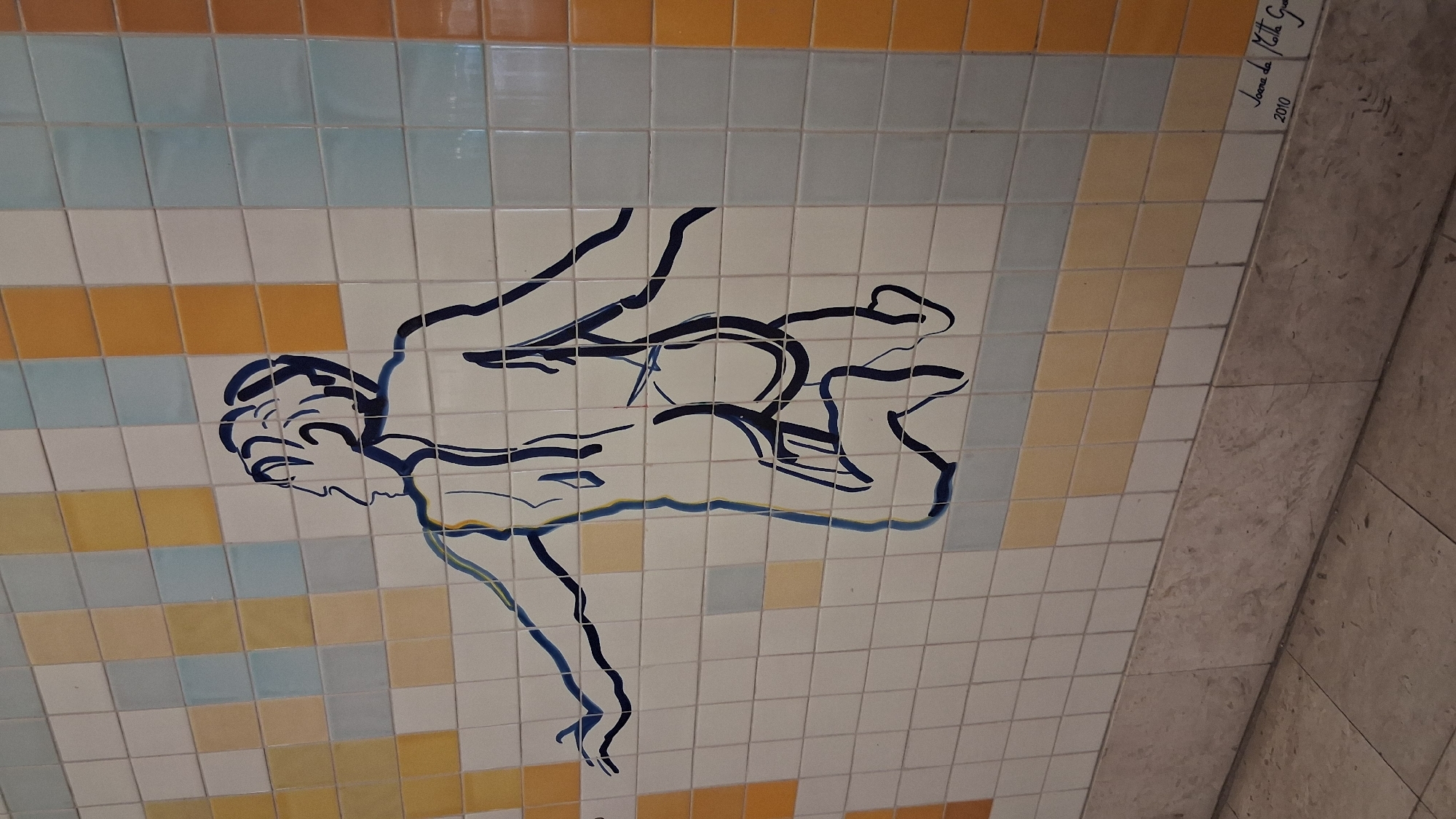 This media file features a close-up, tilted view of a mural created on ceramic tiles. The mural depicts a stylized, minimalist outline of a person in motion, rendered in dark blue ink. The figure appears to be diving or leaping, with limbs extended and body contorted in a dynamic pose. The background is composed of a grid of square tiles in varying colors: white, pale blue, and a muted orange-yellow. The tiles themselves are glossy and reflect light, creating subtle variations in brightness across the image.
Towards the upper right corner of the tiled section, faint, handwritten text is visible in dark ink. The text appears to be a signature and a date, reading "Joana da Motta Gu... 2010".
The tiled mural transitions to a different material on the right side of the frame, which appears to be a rough, textured stone or concrete surface, also laid in a tiled pattern but with larger, irregular rectangular tiles in neutral, earthy tones. This textural contrast suggests the mural is part of a larger architectural installation, possibly in a public space. Given the provided location context of Setúbal, Portugal, this could be a piece of public art within the city. The lighting suggests an indoor setting, possibly a building interior or a covered walkway, with artificial light illuminating the scene. No specific time of day or weather conditions are discernible.
