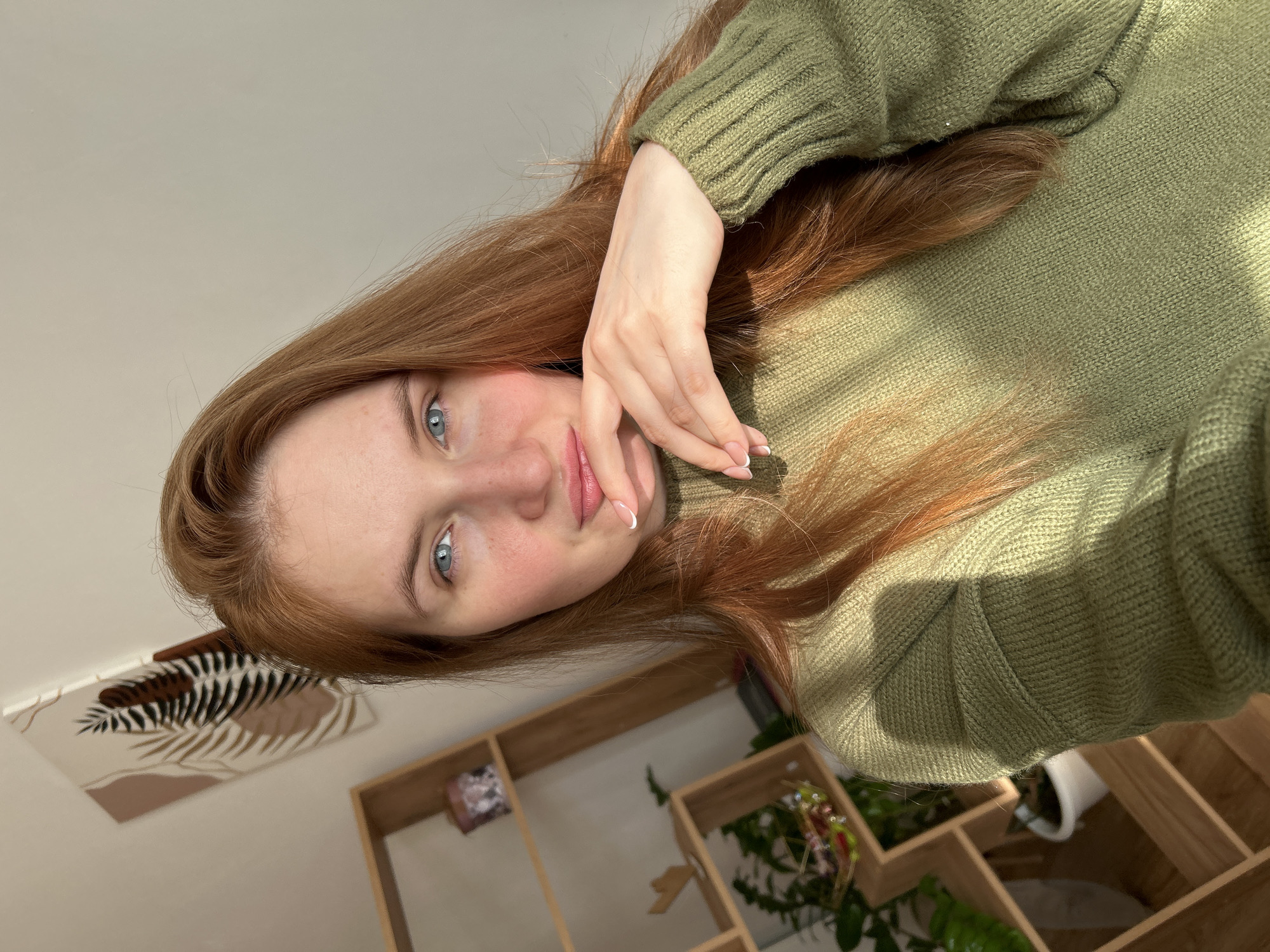 A young woman with long auburn hair and fair skin is facing the camera, her head tilted slightly to the right. Her blue eyes and freckled complexion are visible. She is wearing a green sweater, and her right hand is gently touching her face.
The background includes a wall with a decorative print and a wooden shelving unit containing various items, including plants and decorative objects.
The image was taken indoors, in what appears to be a private residence or apartment. Based on the prompt's location, this is presumed to be in Chaikovsky, Russia.