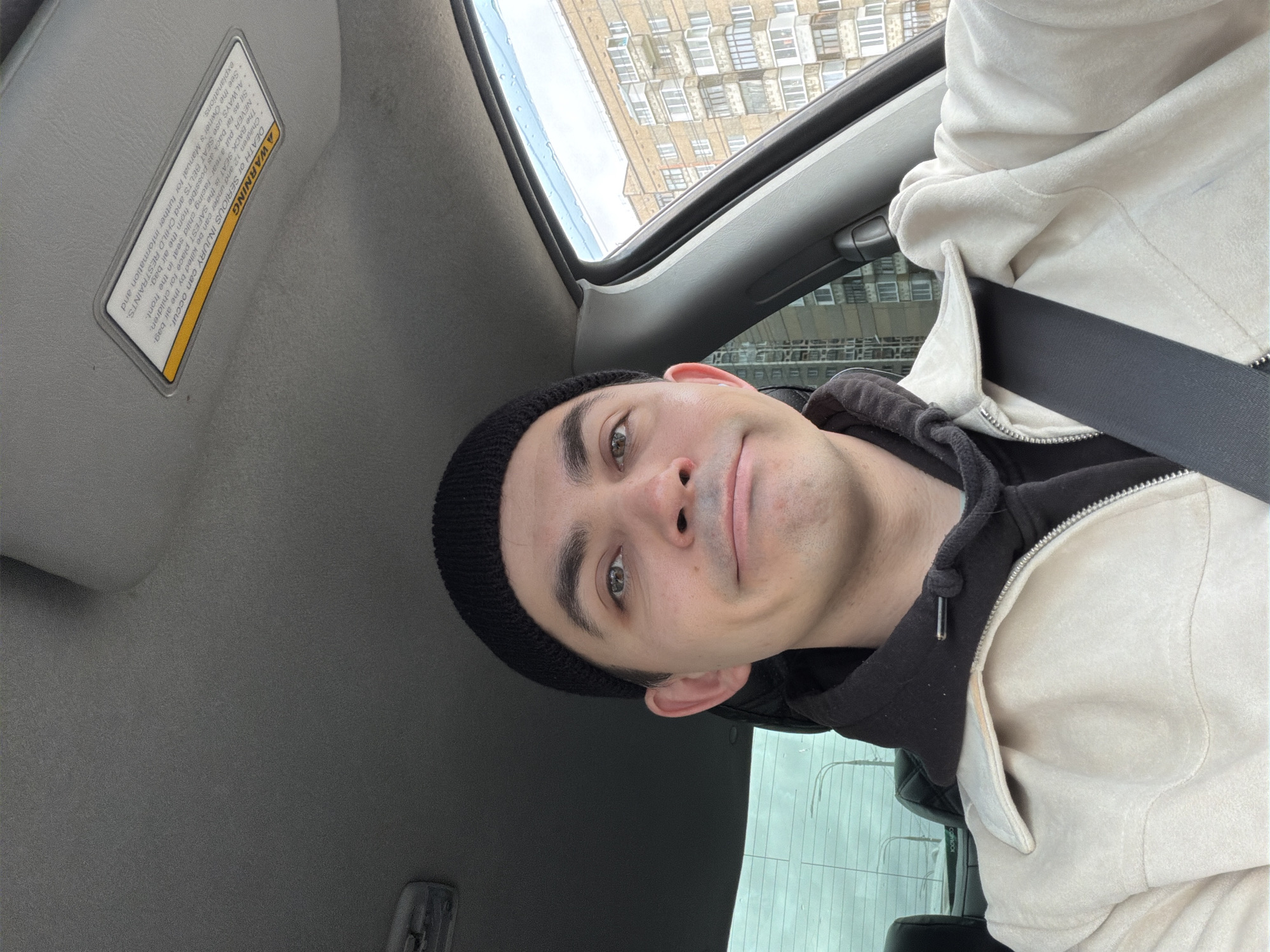 The image presents a selfie of a person wearing a black beanie, a black hoodie, and a light beige jacket. They are positioned inside a vehicle, wearing a seatbelt. Outside the car window, a multi-story building with numerous windows is visible, indicating an urban environment. A sun visor with a warning label is seen on the car's interior. The OCR text includes the word "colmoor." Considering the exterior architecture and the OCR text, it can be assumed that the location could be Izhevsk, Russia.