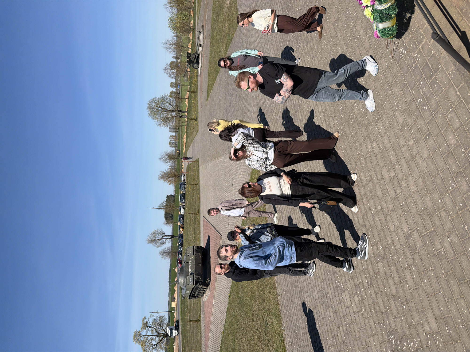 The image depicts a group of eleven people standing on a brick-paved area in the city of Свіслацкі сельскі Савет, Belarus. The people are of various ages and genders, dressed in casual attire. Some are wearing sunglasses. A military vehicle, possibly a tank, is visible in the background to the left. In the background, there are trees and parked cars on the left. On the right of the people, there is a bouquet of flowers. The sky is clear and blue.
