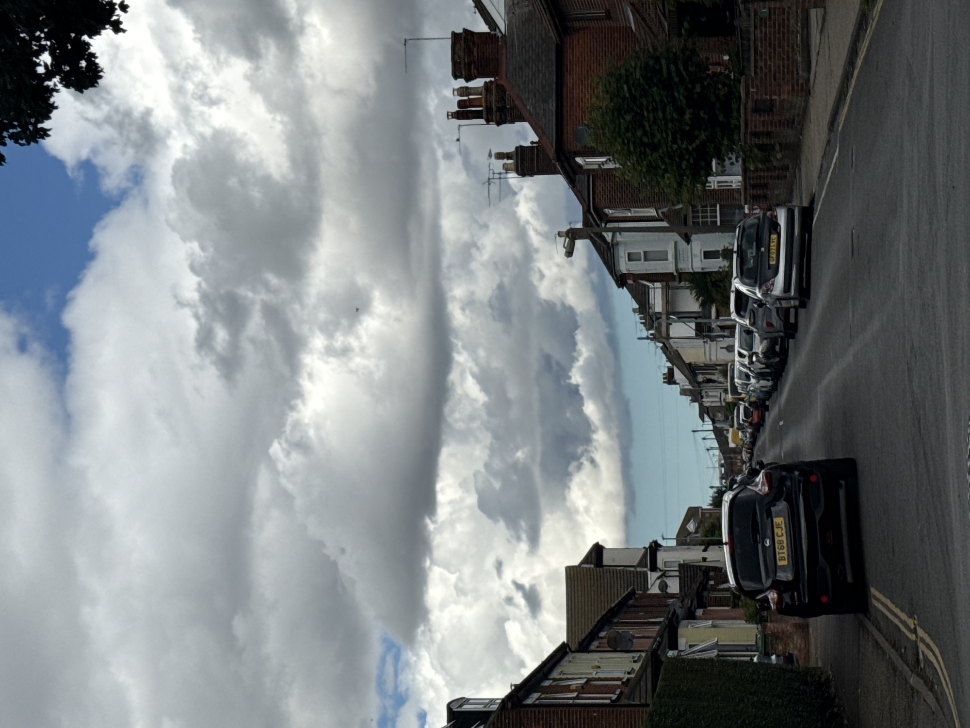 The image depicts a residential street in Great Yarmouth, United Kingdom, under a cloudy sky. The foreground shows a black Nissan Juke with the license plate "BT68 CJE" parked on the left side of the road. The street is lined with terraced houses, primarily constructed of brick, with typical British architectural features such as dormer windows and chimneys. Several other cars are parked along the street, including a silver Peugeot 207 with the license plate "GF07 LVC" further down on the right. The houses have neat gardens and brick walls bordering the sidewalks. The sky is dominated by large, grey, cumulus clouds, with patches of blue visible in the distance. The scene conveys a typical suburban setting in the UK on an overcast day.