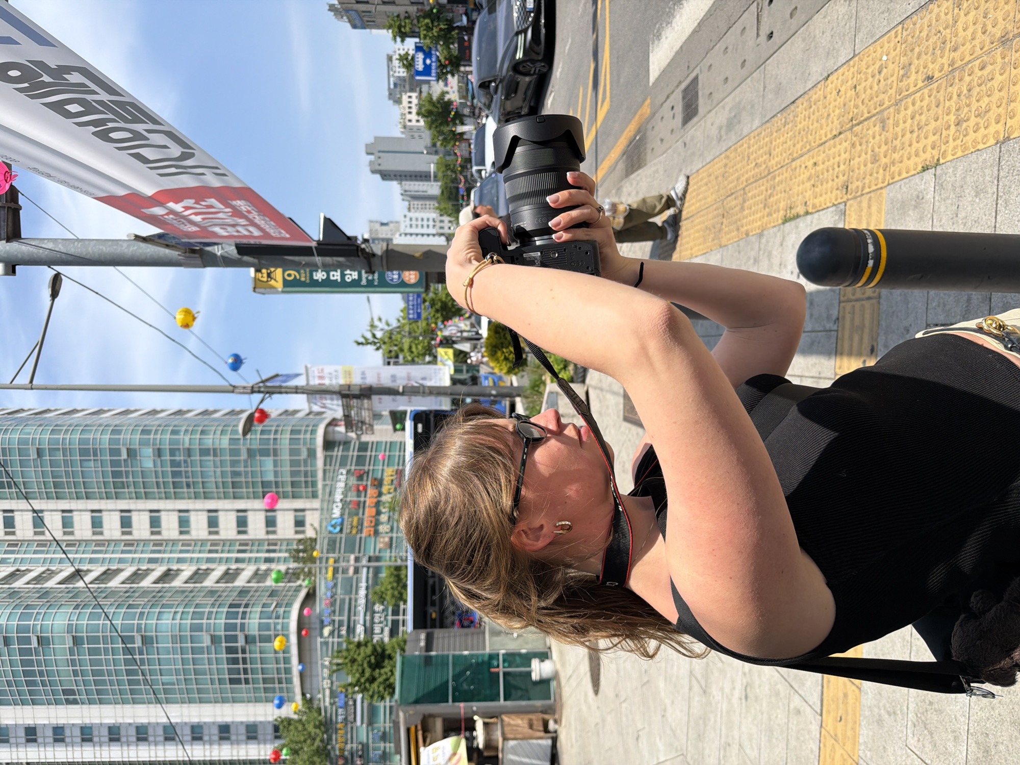 A young woman in a black top and sunglasses is holding a digital camera, pointing it towards a street scene in Seoul, South Korea. She stands on a sidewalk with yellow tactile paving. In the background are modern buildings, street signs with Korean characters, and a large banner also displaying Korean text. Cars are visible on the road. The image is slightly tilted, providing a unique perspective of the urban environment.