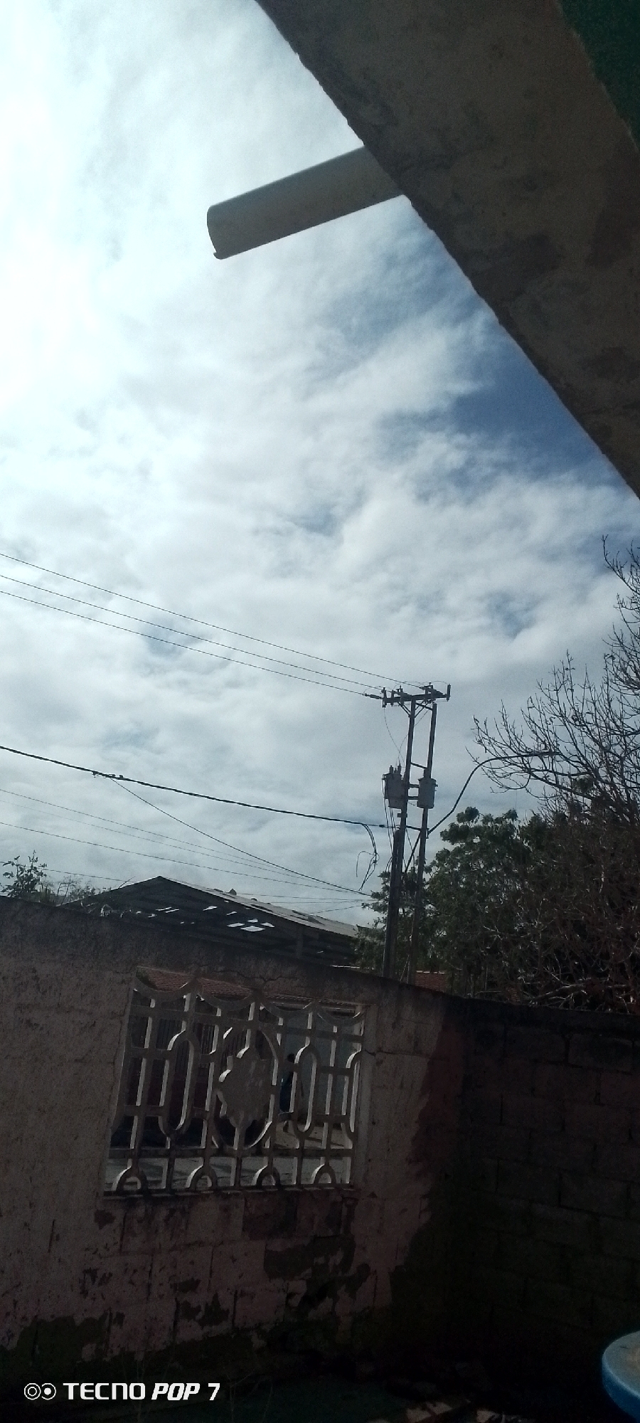 The image shows an exterior scene in Altagracia, Venezuela. A utility pole with transformers is visible against a cloudy sky. Below the pole is a section of a building with a wall and a window with a decorative metal grate. A partially obscured roofline is present behind the wall. Tree branches are visible to the right of the utility pole. The upper portion of the image is partially obscured by a concrete overhang.
