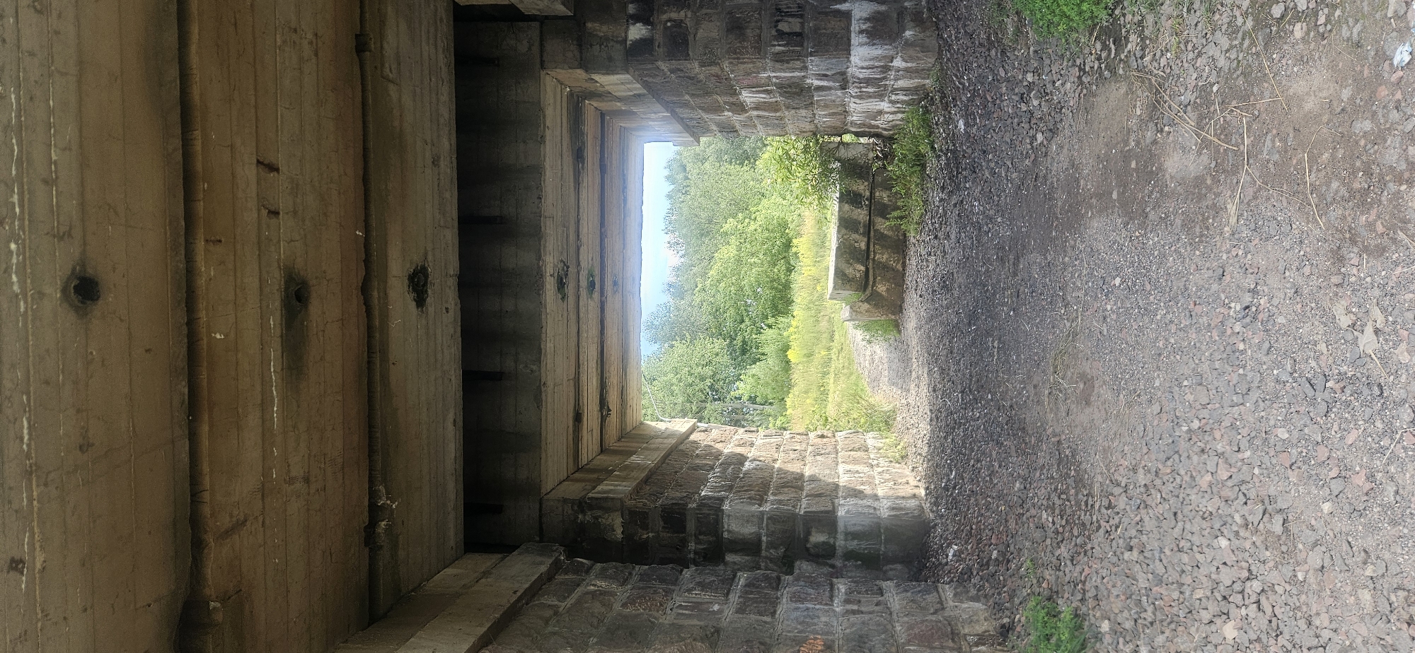 The image shows a view looking through an old concrete and stone bridge structure. The bridge has a rough, weathered appearance, with visible concrete beams and stone block supports. The interior of the bridge is dark, leading to a brightly lit opening. Through this opening, a path made of gravel and dirt leads into a natural environment with lush green trees and foliage under a bright sky. Two large, rectangular concrete blocks are positioned on the right side of the path, near the bridge's exit. The overall scene suggests an abandoned or disused railway bridge in a rural or semi-rural setting, possibly on the outskirts of a city. There are no people or animals visible in the image. The specific location in Vitsebsk, Belarus cannot be determined from the visual information alone.