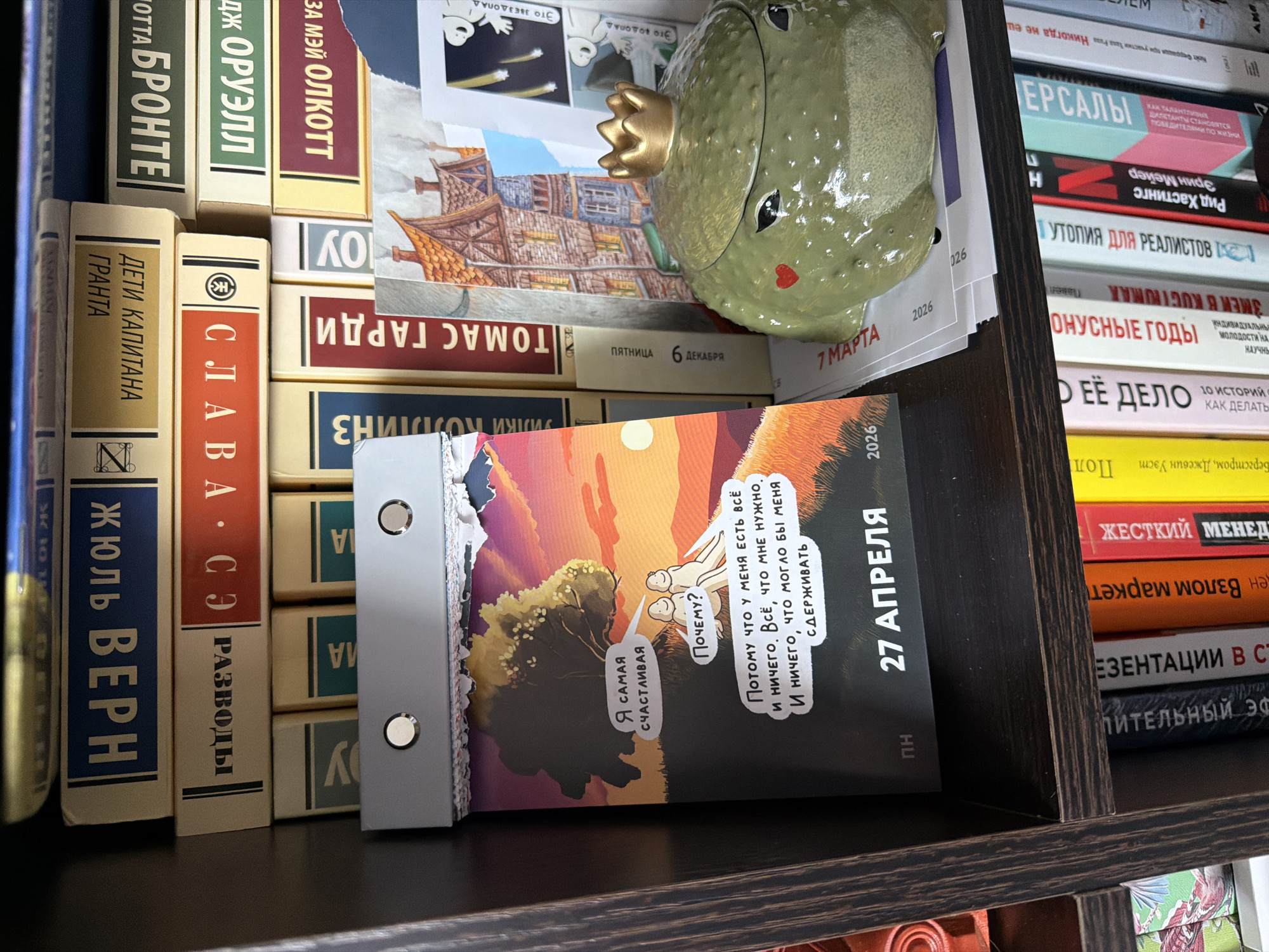 An eye-level shot captures a cluttered but organized section of a dark wooden bookshelf in what appears to be a home setting in Chaikovsky, Russia. The shelf is filled with books and a few decorative items.
On the left, a stack of books, primarily in Russian, stands upright. Visible titles include "ЖЮЛЬ ВЕРН" (Jules Verne) with "ДЕТИ КАПИТАНА ГРАНТА" (Children of Captain Grant), "СЛАВА СЭ" (Slava Se) with "РАЗВОДЫ" (Divorces), "БРОНТЕ" (Brontë), "ДЖ ОРУЭЛЛ" (J. Orwell), and "ЗА МЭЙ ОЛКОТТ" (Z. May Alcott). Another book spine reads "ТОМАС ГАРДИ" (Thomas Hardy).
To the right, a shiny, light green ceramic frog wearing a golden crown sits on top of a horizontal stack of books and papers. The frog has small black eyes and a red heart painted on its cheek. Partially visible under the frog is a card with illustrations and text "ЭТО ЗВЕЗДОПАД" (This is a meteor shower) and "ЭТО ВОДОПАД" (This is a waterfall). Another small card indicates "ПЯТНИЦА 6 ДЕКАБРЯ" (Friday 6 December).
In the foreground, leaning against the horizontal books, is an open desktop calendar or planner. It displays an illustration of two small, white figures under a tree at sunset or sunrise. One figure says "Я САМАЯ СЧАСТЛИВАЯ" (I AM THE HAPPIEST), and the other asks "ПОЧЕМУ?" (WHY?). The response is: "ПОТОМУ ЧТО У МЕНЯ ЕСТЬ ВСЁ И НИЧЕГО. ВСЁ, ЧТО МНЕ НУЖНО. И НИЧЕГО, ЧТО МОГЛО БЫ МЕНЯ СДЕРЖИВАТЬ" (BECAUSE I HAVE EVERYTHING AND NOTHING. EVERYTHING I NEED. AND NOTHING THAT COULD HOLD ME BACK). The visible date on this calendar is "27 АПРЕЛЯ" (27 April) 2026, marked as "ПН" (Monday). Another card behind the frog shows "7 МАРТА 2026" (7 March 2026).
Further to the right, another section of the bookshelf holds more upright books. Titles here include "ЕРСАЛЫ" (likely Jerusalem), "РИД ХАСТИНГС" (Reed Hastings), "УТОПИЯ ДЛЯ РЕАЛИСТОВ" (Utopia for Realists), "БОНУСНЫЕ ГОДЫ" (Bonus Years), "О ЕЁ ДЕЛО" (About Her Business), and "ЖЕСТКИЙ МЕНЕД..." (Harsh Manag..., likely Management). The items suggest a collection of literature, self-help, and business books, interspersed with personal mementos.