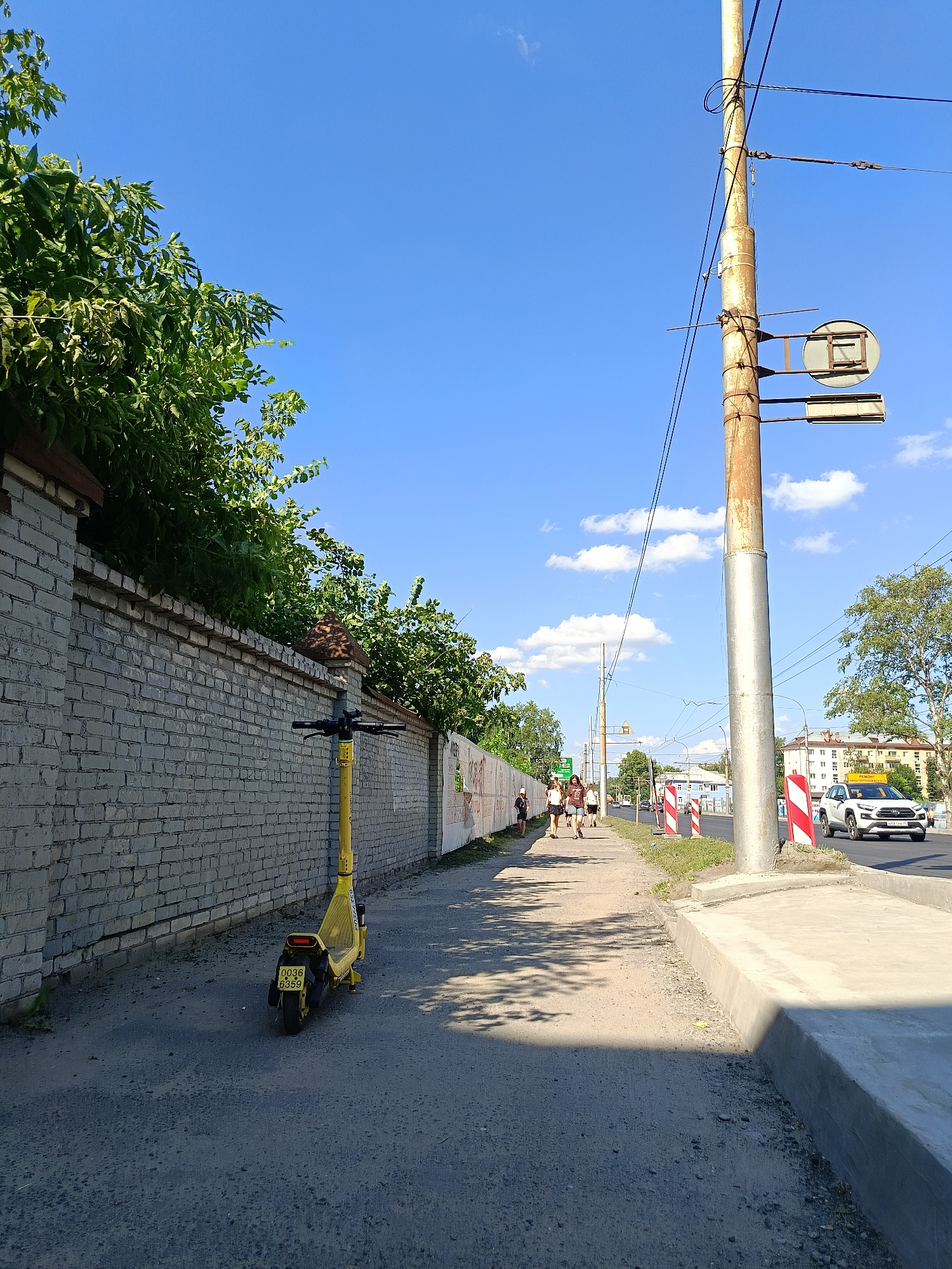 A bright, sunny day in an urban environment, likely a sidewalk next to a road. A yellow electric scooter is parked on a dirt and gravel path. The path runs alongside a light grey brick wall covered in green foliage. Further down the path, several people are walking. On the right side of the image, a utility pole stands tall, supporting wires and signage. A white SUV is visible driving on the road next to the sidewalk, which is bordered by a concrete curb and grass. Buildings are visible in the background.