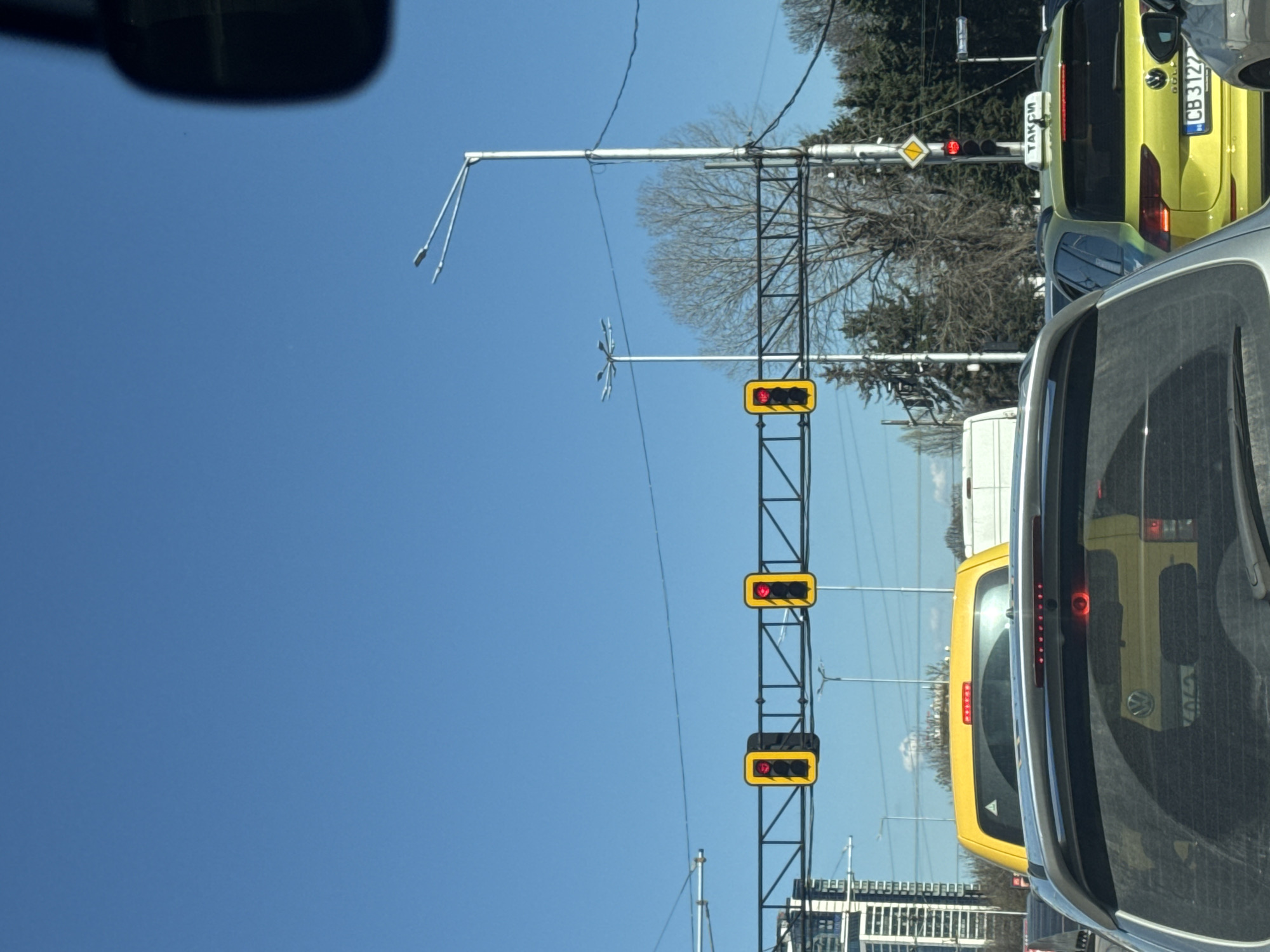 The image shows a street scene in Sofia, Bulgaria. The sky is blue, and there is a black metal traffic light pole with three red lights. The top and bottom lights are rectangular, while the middle is round. A yellow diamond-shaped sign is above the top light. A yellow "Taxi" vehicle is visible, as is a silver car, which appears to be the source of the image. The license plate on the taxi vehicle is "CB3122." Bare trees and buildings can be seen in the background.