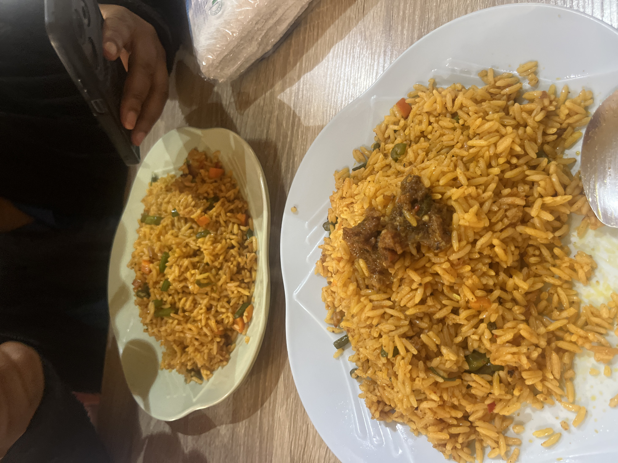 Two plates of Jollof rice are presented on a wooden table. The plate in the foreground, a white ceramic dish, contains a generous portion of Jollof rice topped with a dollop of stew. In the background, a lighter colored plate also holds Jollof rice. A person's hands are visible in the upper left and right quadrants of the image, with the hand on the right holding a smartphone. To the right of the background plate, a stack of wrapped napkins is present. The lighting suggests an indoor setting, possibly a restaurant. The scene is set in Jos, Nigeria.