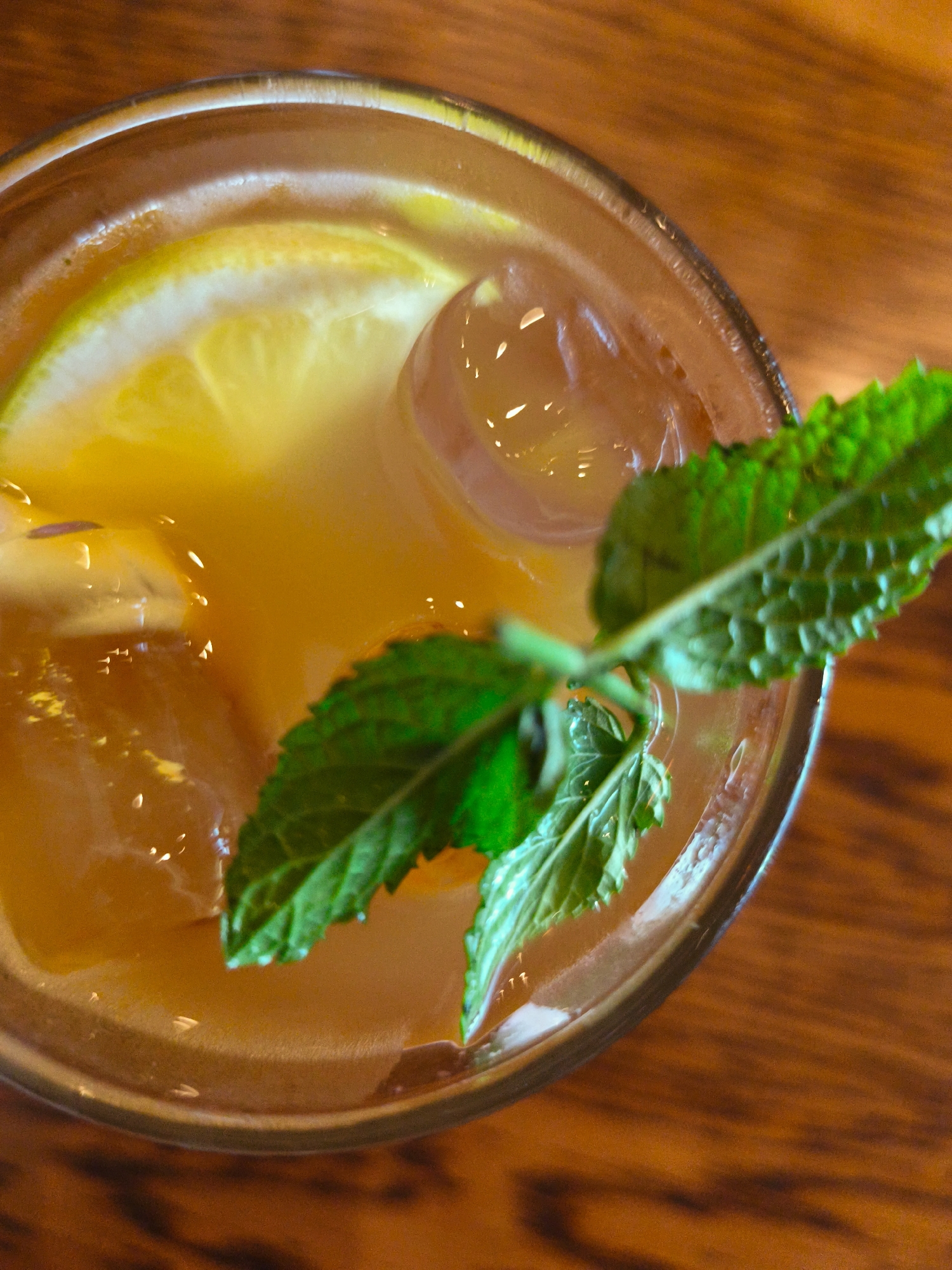 An overhead, close-up shot captures a glass of iced tea with lemon and mint, set on a wooden surface. The drink is a warm amber color, with a slice of lemon and several ice cubes visible within. A sprig of fresh mint rests on top of the ice and liquid, its green leaves contrasting with the beverage. The background consists of a textured wooden table, suggesting an outdoor or casual indoor setting. The image does not contain any identifiable landmarks or elements that would indicate its specific location within Be'er Sheva, Israel.