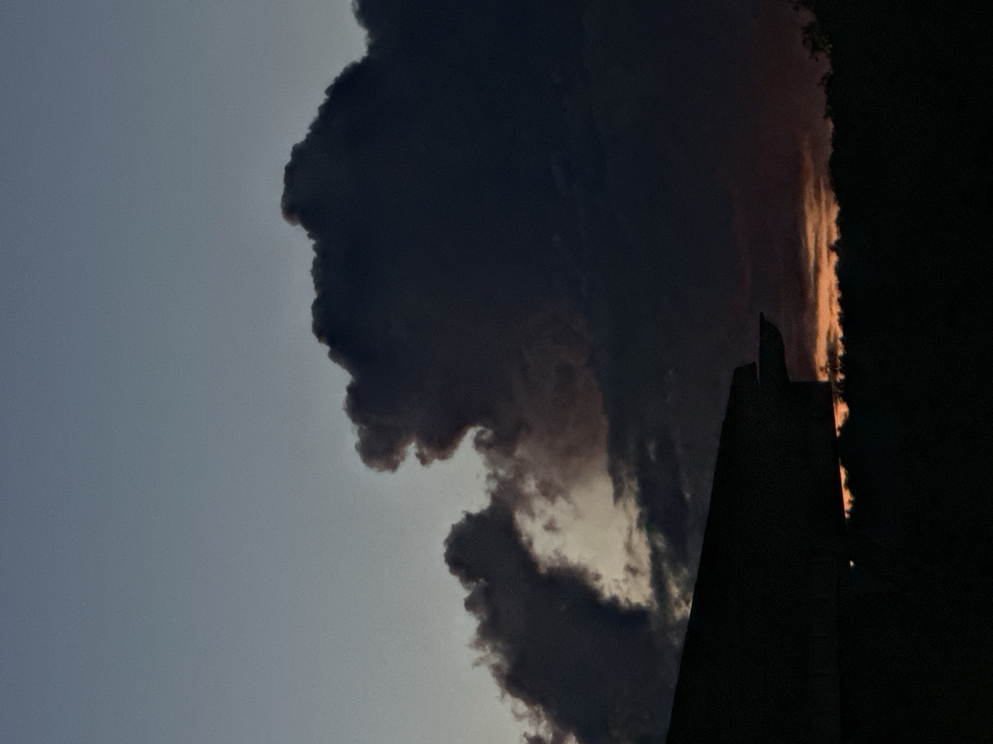 A dimly lit, silhouetted building's roofline occupies the lower left portion of the frame, casting a shadow over a dark, brush-filled landscape. Above the building, large, dark clouds dominate the sky, their ominous forms partially illuminated by a soft, warm glow from the horizon. The glow, hinting at a sunset or sunrise, is tinged with orange and red, creating a stark contrast with the deep blues and grays of the upper sky and the heavy clouds. The overall atmosphere is somber and dramatic, with the subdued lighting and imposing cloud formations suggesting a twilight or stormy evening. There are no discernible people, animals, or specific landmarks that would allow for pinpointing the exact location within Nizhny Novgorod, Russia, based solely on this image.