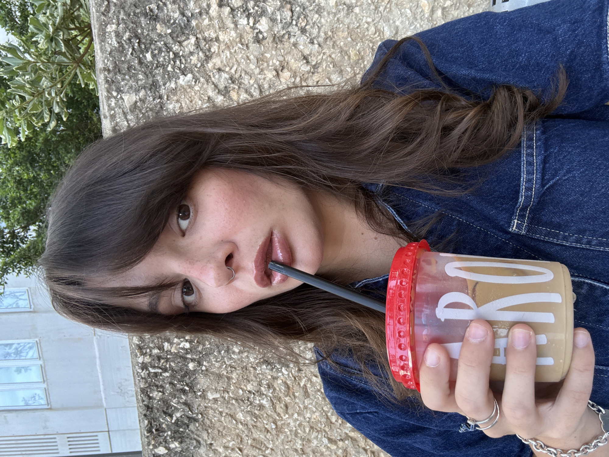A close-up, waist-up portrait of a young woman with dark brown hair and bangs is shown. She is wearing a denim jacket and holding a clear plastic cup with a red lid, containing a light-colored beverage and a black straw. She is looking towards the camera and appears to be sipping from the straw. The background consists of a textured stone wall and some greenery, suggesting an outdoor setting. The overall lighting is natural and appears to be daylight. There is no indication of the specific location within Tel-Aviv, Israel, but the style of architecture and vegetation could be consistent with urban outdoor spaces in the city. The image focuses on the woman and her drink.