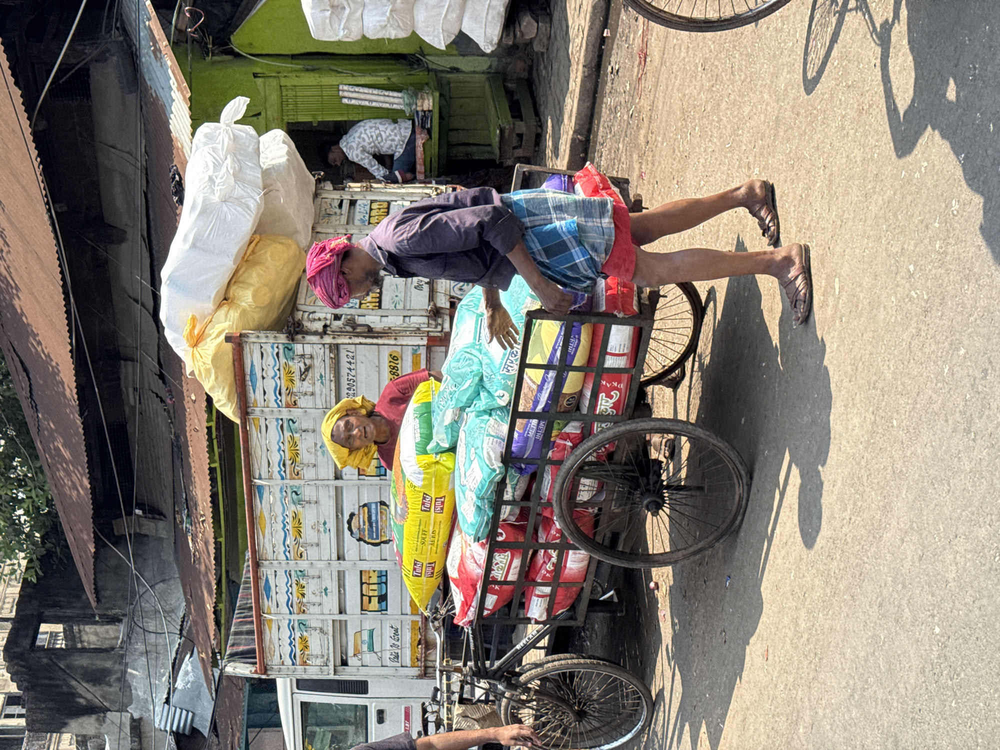 The scene depicts an outdoor urban street in Kolkata, India, under strong daylight. In the foreground, a man wearing a pink turban, a dark blue collared shirt, and a blue-and-white checkered lungi over red shorts, with brown sandals, is positioned next to a black, three-wheeled bicycle-cart. He appears to be handling the numerous multi-colored sacks (yellow, red, light blue, purple) loaded onto the cart, some visibly labeled "Tulsi Souff" and "METI." Behind the bicycle-cart, a woman with a yellow head covering and a red shirt is partially visible. To the far left, the arm and side of a person wearing a dark shirt are seen.
Behind the central activity, a white commercial truck with colorful decorative painting is parked, its open bed piled with large yellow and white sacks. The truck's rear displays Bengali script identifying 'Kolkata' (কলকাতা) and the number '8816'. To the right of the truck, a bright green-painted shop or stall with a corrugated metal roof is visible. Inside this shop, a man in a light patterned shirt is seated on a wooden bench. Stacks of white sacks are present outside the green shop. The background consists of older, multi-story buildings with discolored, weathered facades and corrugated iron roofs. Overhead electrical wires crisscross the upper portion of the scene. The street surface is paved, appearing dusty and containing minor debris, with long shadows cast by the objects and people.