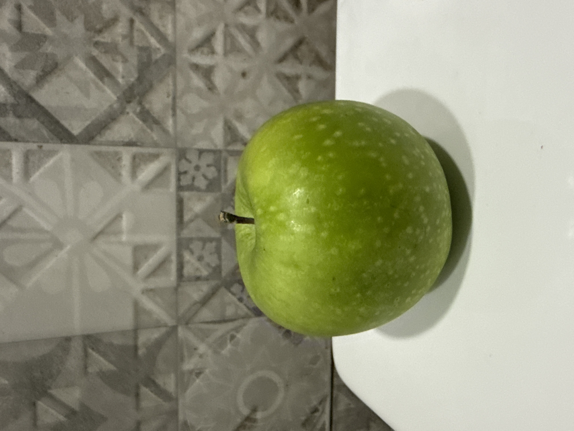 A single, bright green apple with small white speckles sits on a white surface. The apple has a short, brown stem. Behind the apple is a wall covered in decorative tiles with a geometric pattern in shades of grey and white. The lighting suggests an indoor setting, possibly a kitchen or dining area. The image does not provide any specific details about the location within Tbilisi, Georgia.