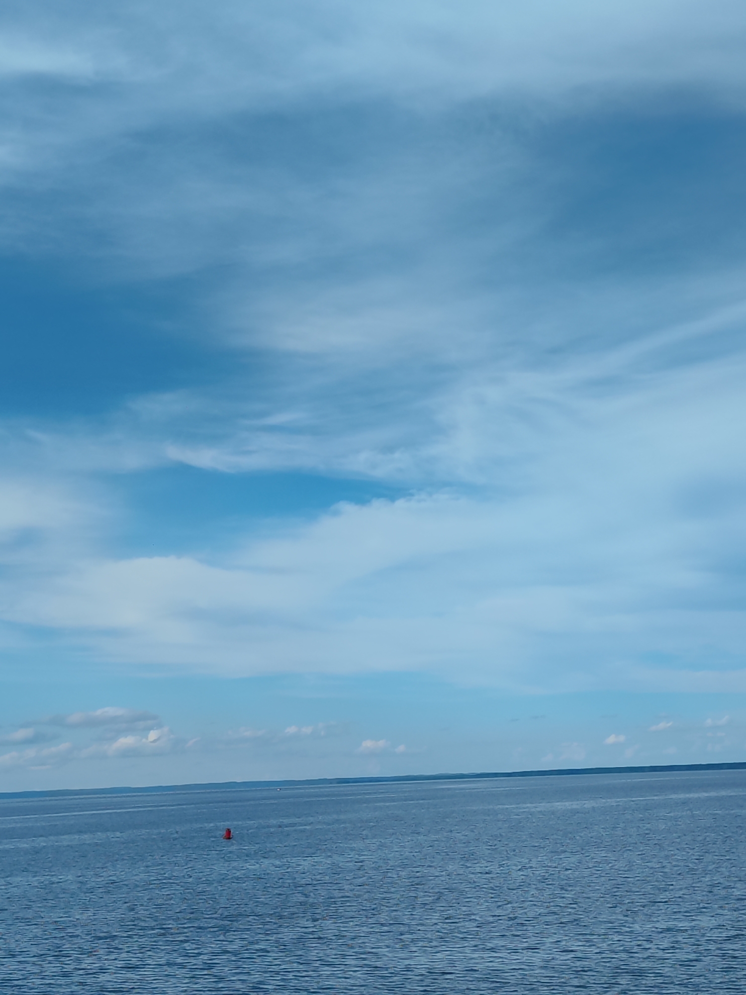 An eye-level, wide shot captures a vast body of tranquil blue water under a sky filled with wispy white clouds. In the distance, a single, small red buoy floats on the water's surface. The horizon line, where the water meets the sky, is sharp and dark, suggesting land in the far distance. The water is calm, with gentle ripples disturbing its surface. The overall scene evokes a sense of peace and openness.