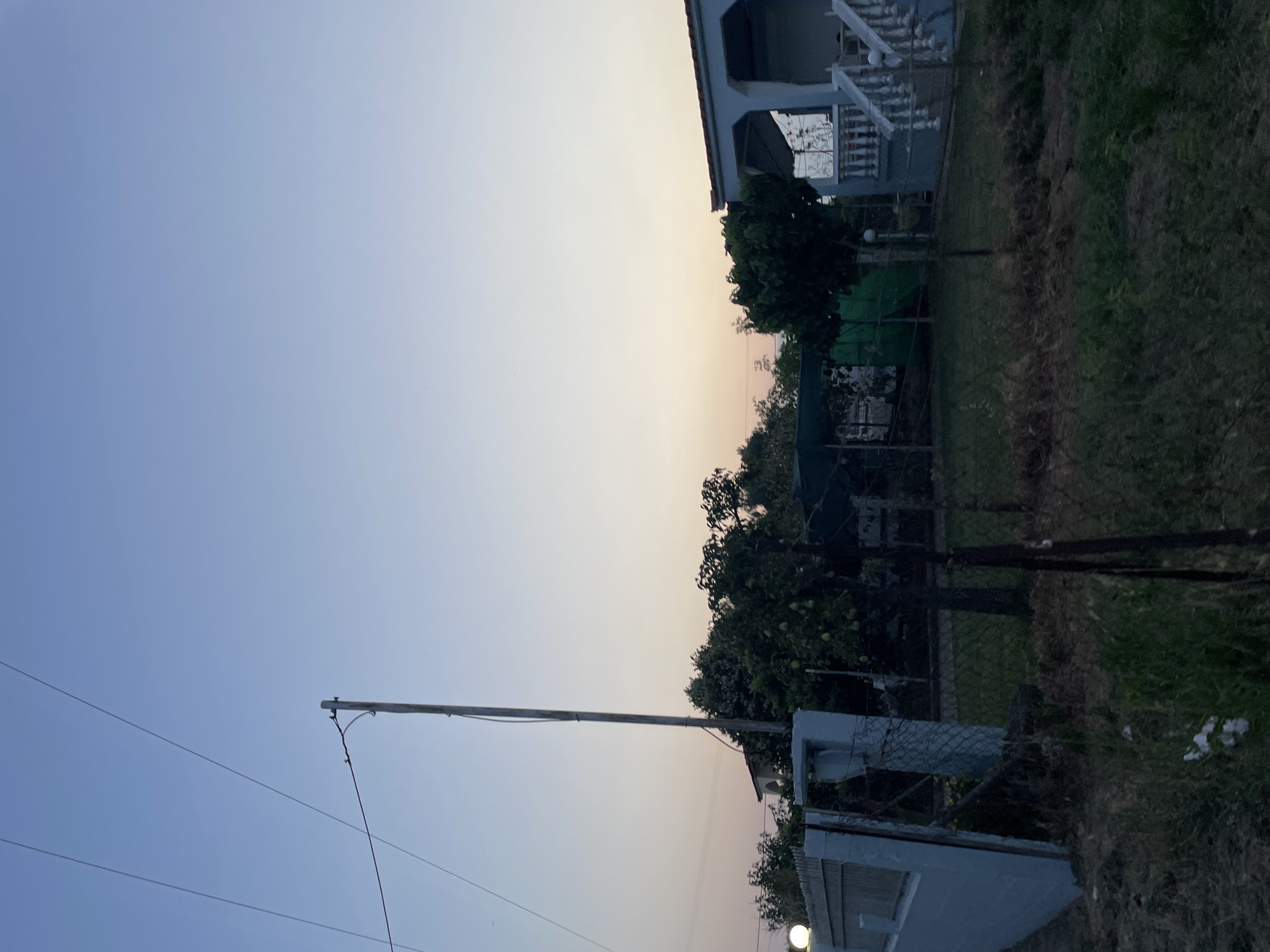 The image depicts a residential scene at dusk in Skala Avdiron, Greece. The sky is a gradient of soft blues and pale oranges, indicative of twilight. A wooden utility pole stands prominently on the left side of the frame, supporting power lines that stretch across the sky. In the foreground, a light blue wall or fence extends from the left edge of the image. Adjacent to this wall is a wire fence. Beyond the fence, a grassy yard is visible, with several trees and some covered structures. On the right side of the image, a white-columned house with a porch and a staircase is partially visible. The overall impression is a quiet, rural setting as day transitions into night.