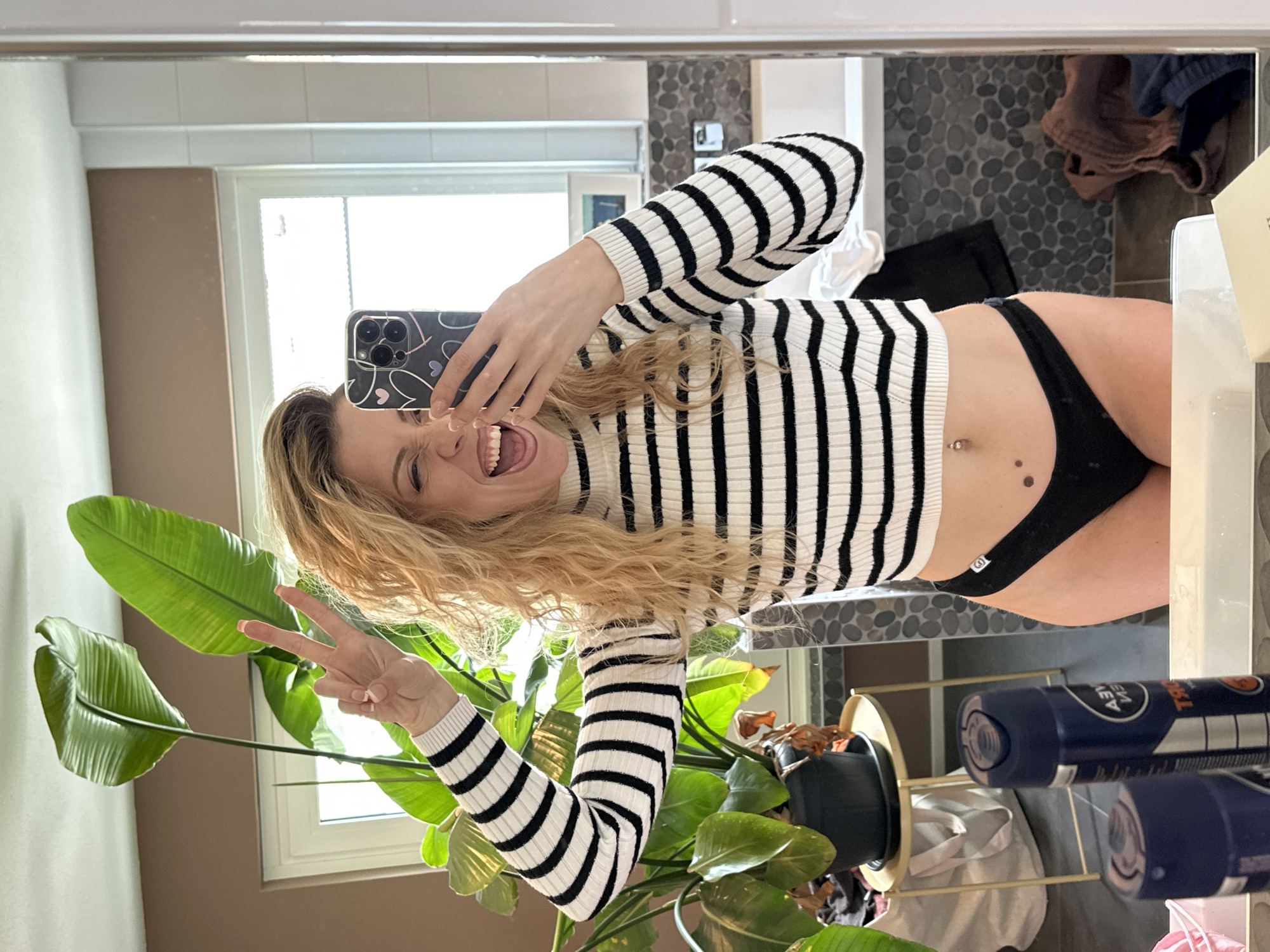 The image is a selfie taken in a bathroom, likely in Dronten, Netherlands, given the prompt. The subject, a blonde woman, is holding a phone up to the mirror, displaying a peace sign. She is wearing a black and white striped turtleneck and black underwear. A plant stands to her left, next to a window. The background of the mirror shows a tiled wall, clothes, toiletries, and what appears to be a sink area.