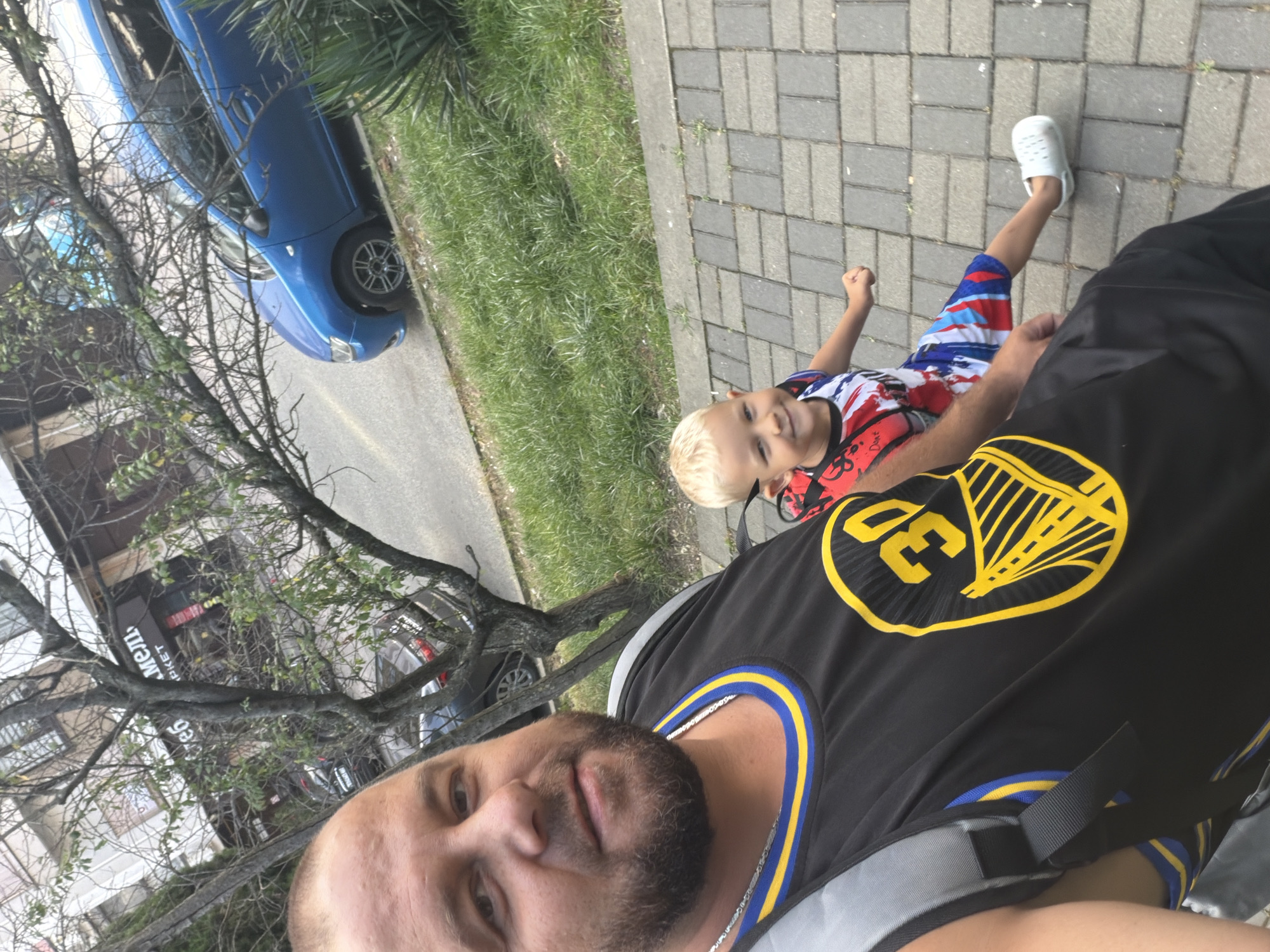 The image depicts a father and his young son walking on a sidewalk in Sochi, Russia. The father, who is in the foreground, is wearing a black basketball jersey with the number "30" in yellow, and a silver chain. He has a beard and is looking towards the camera. His son, to his right, is wearing a red, white, and blue patterned t-shirt and white crocs, and is smiling as he walks. The background shows a street with a blue car parked on the side, and a building with a sign that reads "МЕЛЬ" and "МАРКЕТ" in Cyrillic. A tree with bare branches is also visible, suggesting it might be autumn or winter. The sidewalk is paved with gray and beige square tiles, and there is green grass on the side of the street. The overall scene appears to be a casual outing in an urban environment.