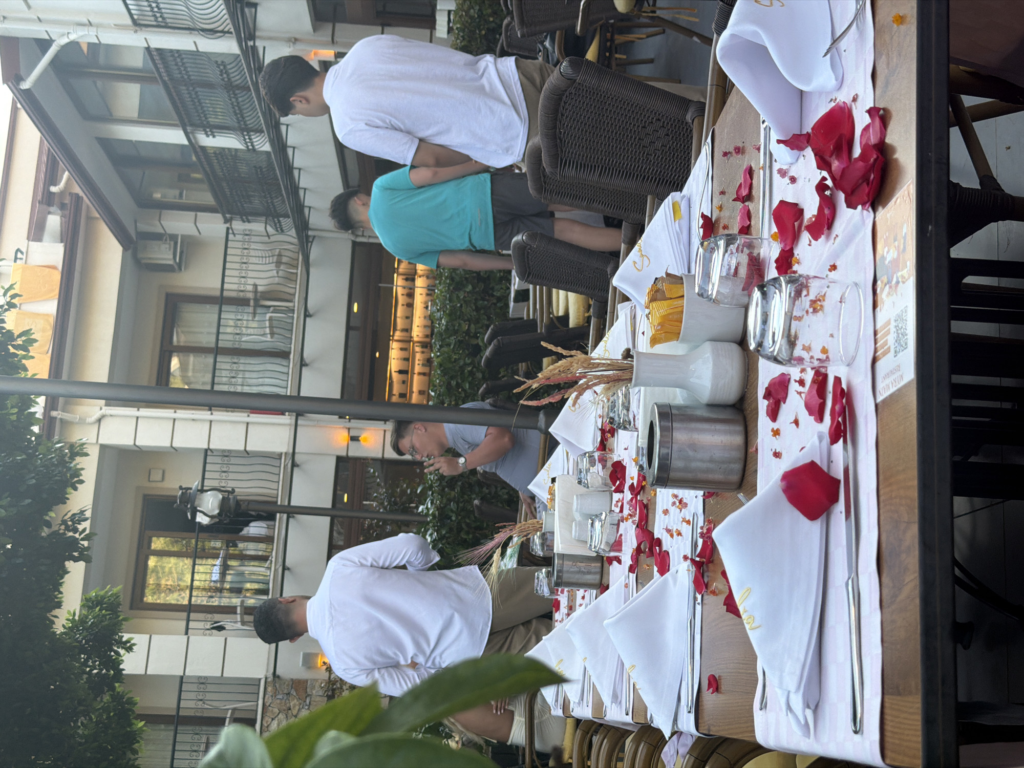 This image captures an outdoor dining scene at what appears to be the "MYRA MAIN RESTAURANT" in Fethiye, Turkey, as indicated by visible text on a table card. The setting features several wooden tables elaborately set with white tablecloths, white cloth napkins embroidered with gold text (possibly "Syra"), silverware (knives and forks), and drinking glasses. Red rose petals and some smaller yellow/orange petals are scattered decoratively across the tables and inside some glasses, suggesting a romantic or celebratory occasion. Small silver containers and white dispensers are also present on the tables.
Several men are visible in the scene. One man in a white long-sleeved shirt and light brown pants stands near the foreground, facing away from the viewer. Another man in a white short-sleeved t-shirt and dark shorts stands further back, also facing away. A man in a teal short-sleeved t-shirt and dark shorts is seen in the mid-ground, viewed from behind. Towards the center of the image, a man wearing a light blue t-shirt and glasses is seen from the side, leaning over one of the tables, possibly interacting with an object on it. Another man in a white long-sleeved shirt is partially visible in the background. The individuals appear to be guests or staff members standing or milling around the dining area.
The background reveals multi-story buildings with balconies featuring intricate wrought-iron railings, consistent with local architecture. Air conditioning units are visible on some buildings. Lush green trees and bushes are scattered around the outdoor space, adding to the natural ambiance. The lighting suggests it is daytime, possibly late afternoon or early evening, with soft, natural light illuminating the scene. The overall impression is one of a pleasant, elegant outdoor dining experience. Yellow sugar packets are also discernible on some tables.