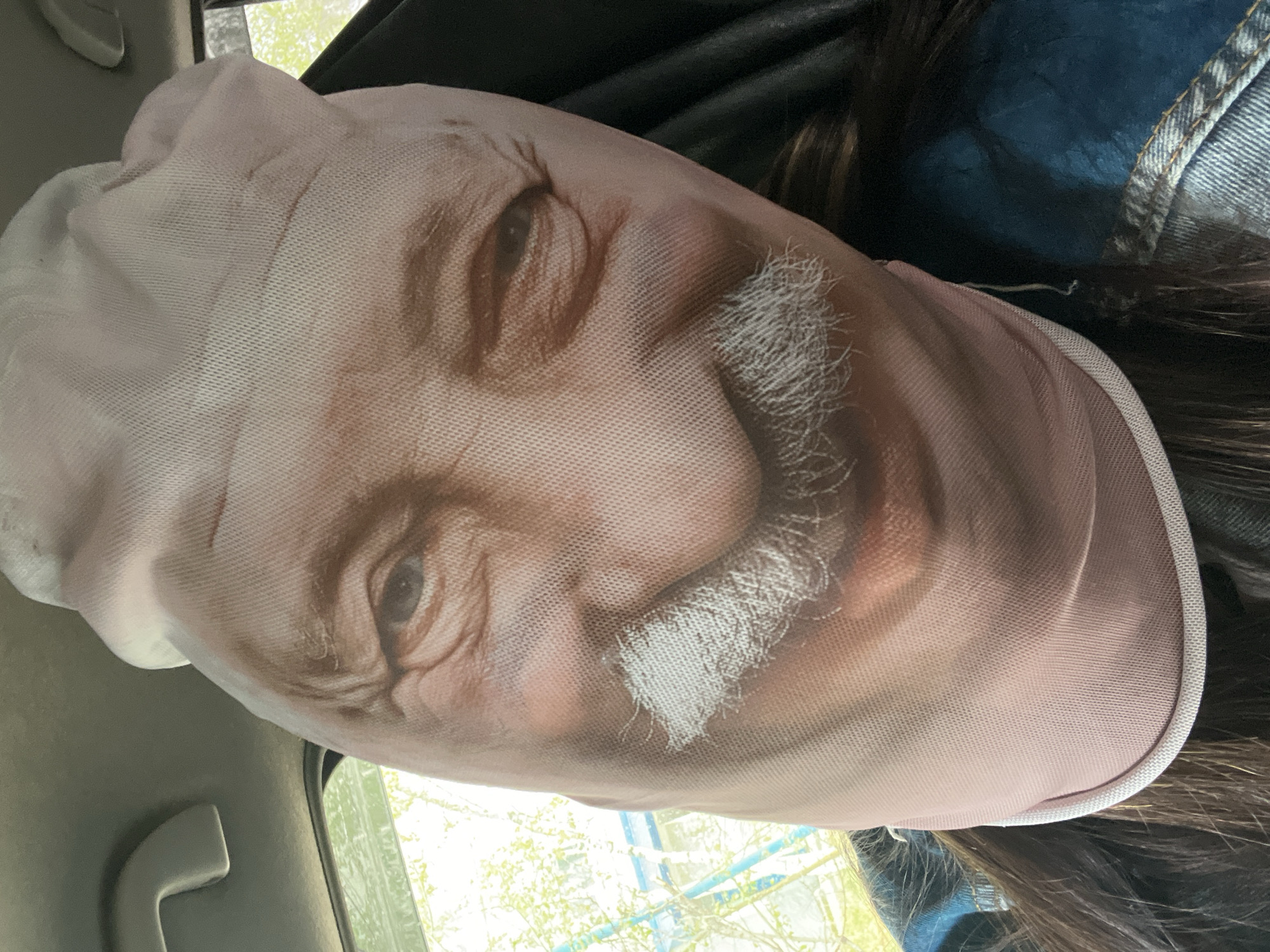 A person wears a mask printed with the face of an elderly man with a white mustache. The person is inside a vehicle, as evidenced by the car door and window visible in the background. The background shows greenery suggesting the vehicle might be outdoors. The location is Unknown, Unknown. The person is wearing a denim jacket. The lighting suggests daytime.
