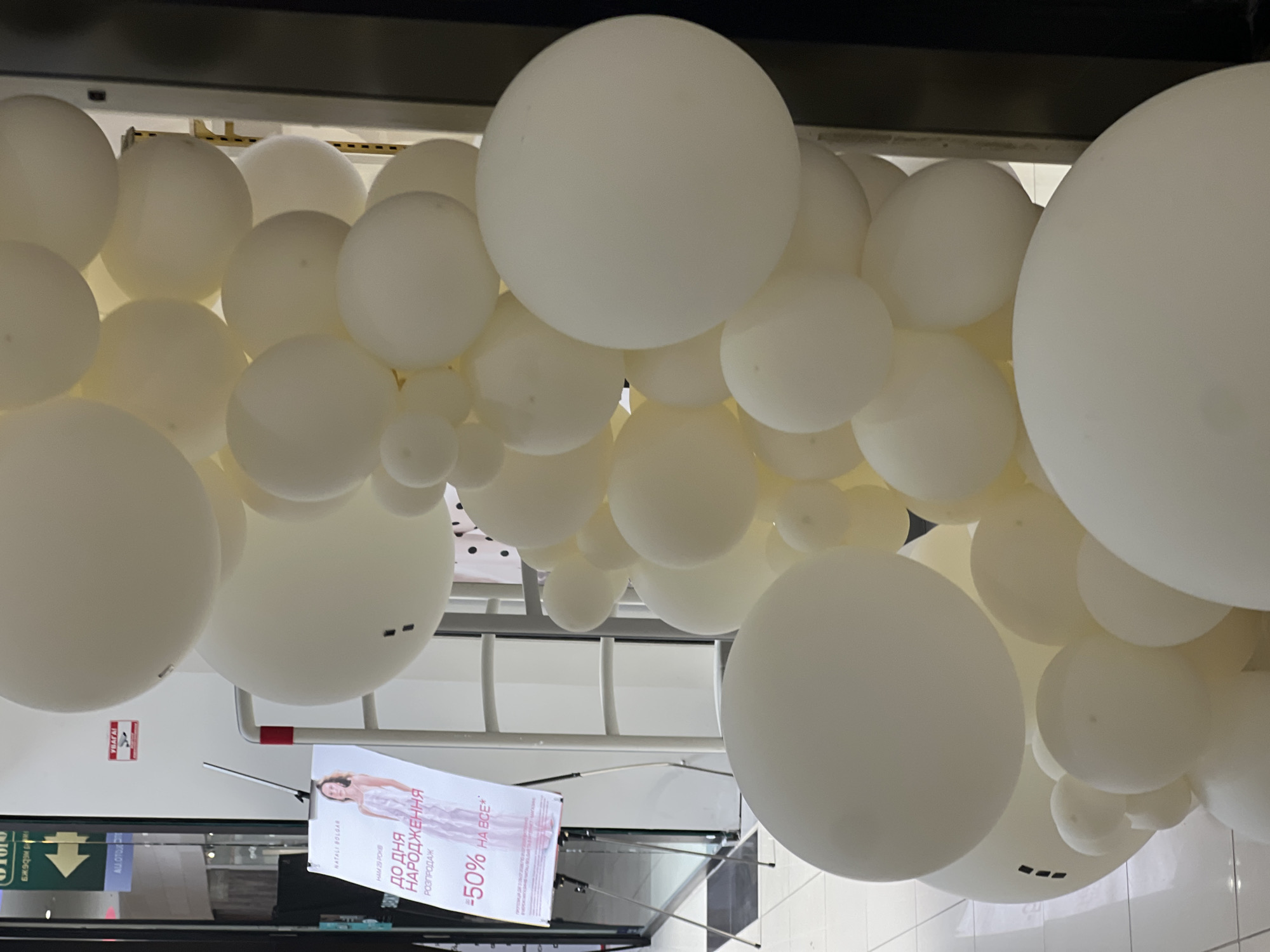 This is a vertical, medium-shot photograph of a balloon arch at an indoor retail location in the city of L'isky, Ukraine. The balloons are uniformly white. The arch is positioned to the right of the frame, with a promotional sign to the left. The sign displays a woman in a white dress and text that reads "НАРОДЖЕННЯ" (Birth) and "-50% НА ВСЕ" (-50% ON EVERYTHING). The sign is supported by a silver tripod stand. In the background, a white metal rack with horizontal bars is visible, suggesting a store fixture. Behind the rack, a blurred storefront or interior is visible, featuring signage with Ukrainian text. A security camera and a fire alarm are mounted on the wall above and to the left of the sign. The floor is tiled in a light color.