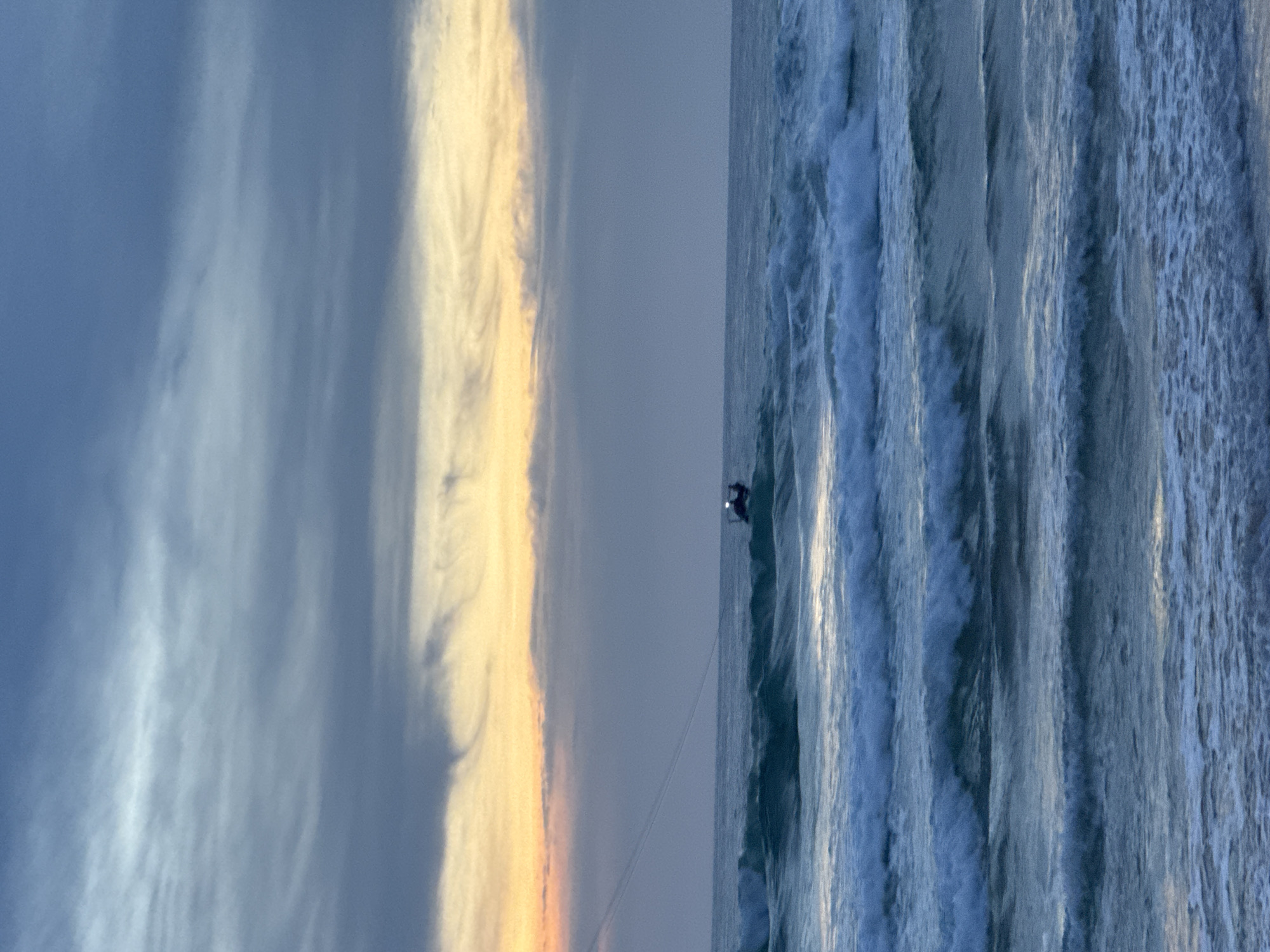 The image shows a body of water with waves breaking on a beach. The sky is overcast with streaks of yellow and orange, suggesting a sunset or sunrise. In the distance, a small boat with a light on is visible on the water. The water appears to be a deep blue-green, and the waves are white-capped. The overall impression is one of a tranquil, natural scene. The location is not specified in the image.