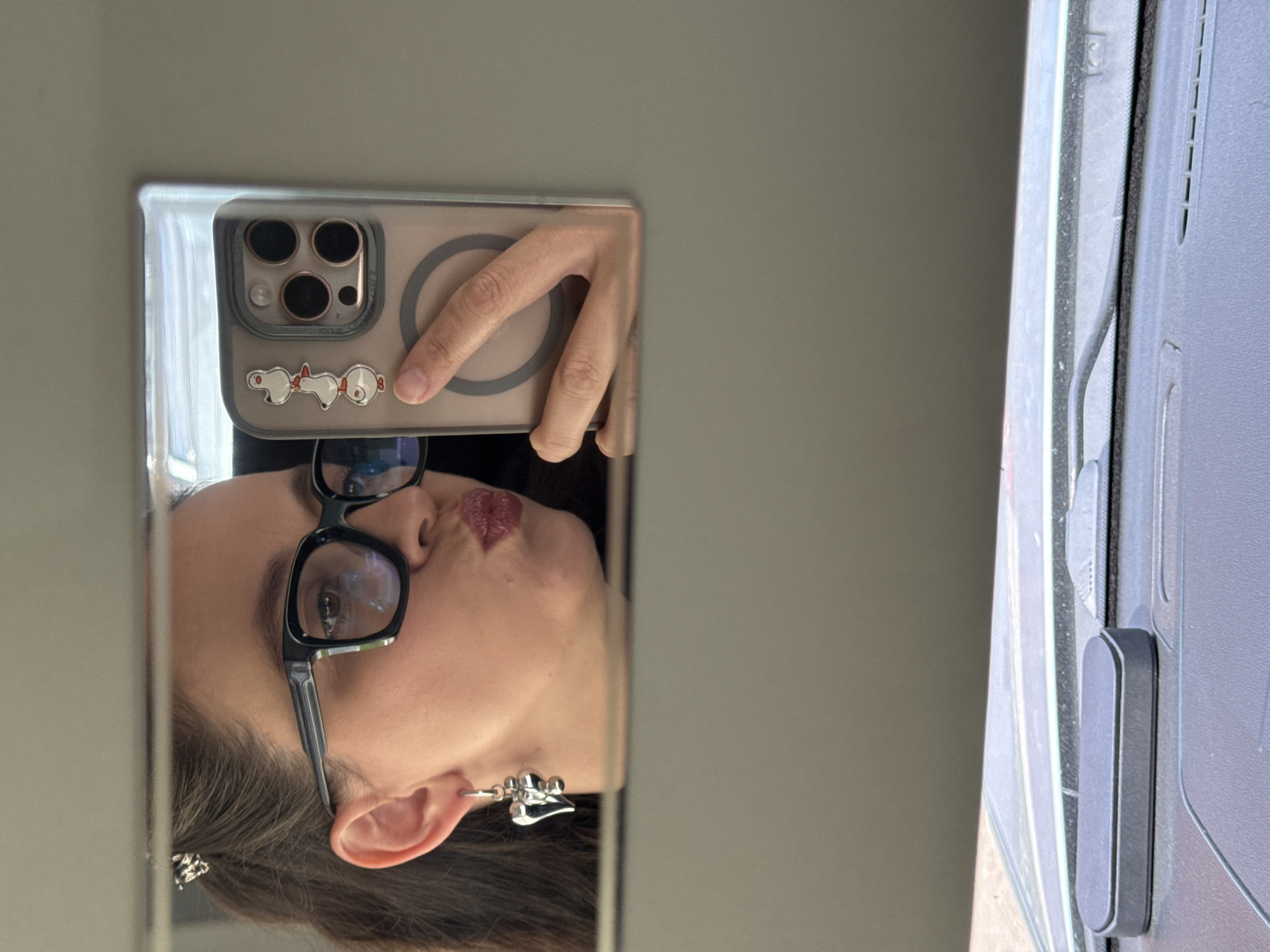 A woman is taking a mirror selfie in a car. She is wearing large black-rimmed glasses and a silver heart-shaped earring. Her hair is pulled back with a small silver clip. She has on a light pink lipstick and is puckering her lips. She is holding a clear phone case with three duck stickers on it. The background shows the interior of a car, including the dashboard and windshield. The image appears to have been taken indoors, possibly in a parking garage, or on a cloudy day, as there is no direct sunlight visible. The location is not specified as being in the city of Chaikovsky, Russia.