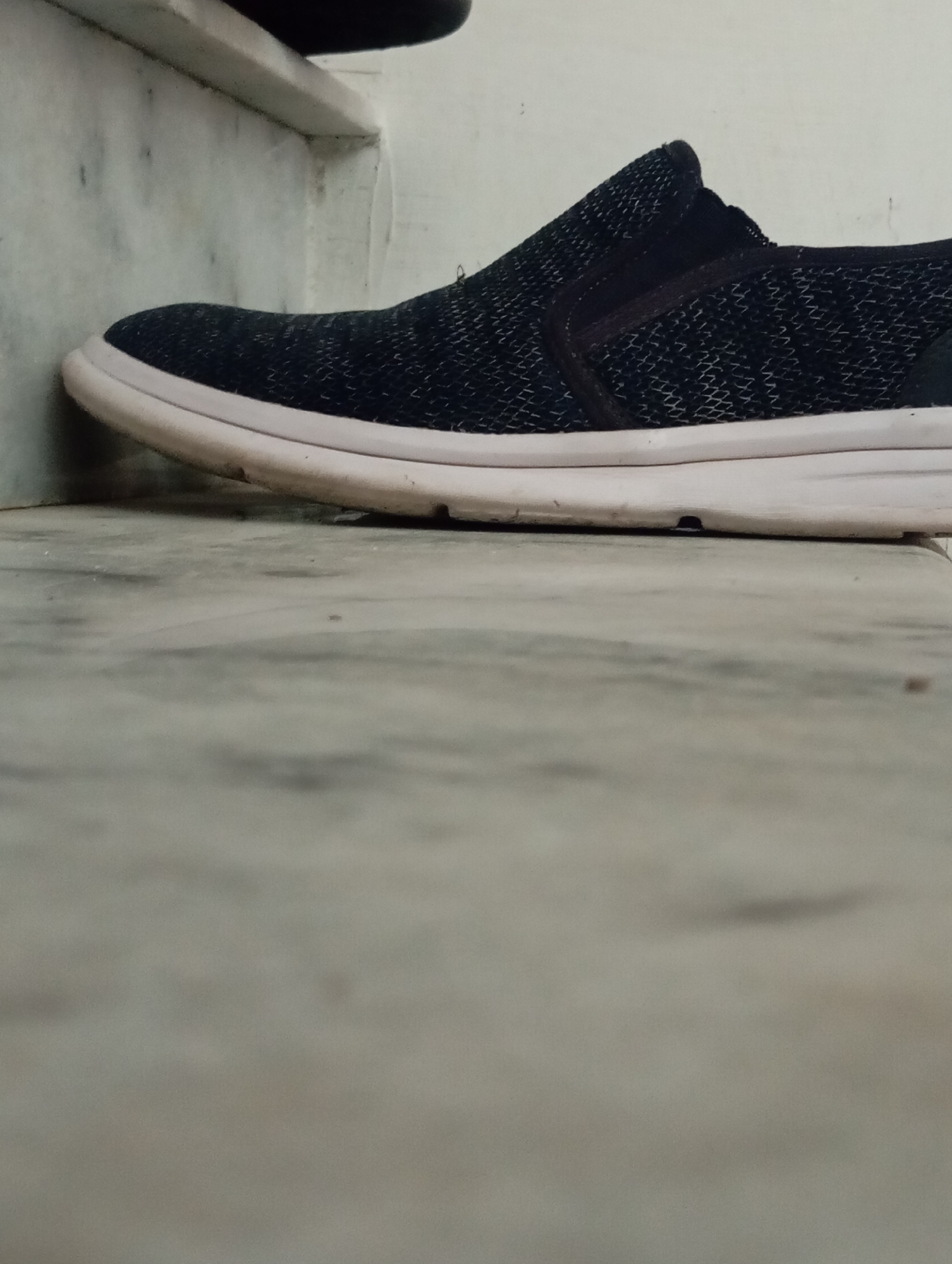 A single dark blue slip-on shoe with a white sole is placed on a marble floor. The shoe is a close-up shot, with the toe of the shoe leaning against a white wall or ledge. The shoe has a textured knit pattern and elastic goring on the sides. The white sole shows some dirt and wear. The background is blurred, but appears to be a marble surface with some shadows. The lighting is somewhat dim, suggesting it may be indoors or late in the day. No people are visible, and no text is present in the image.