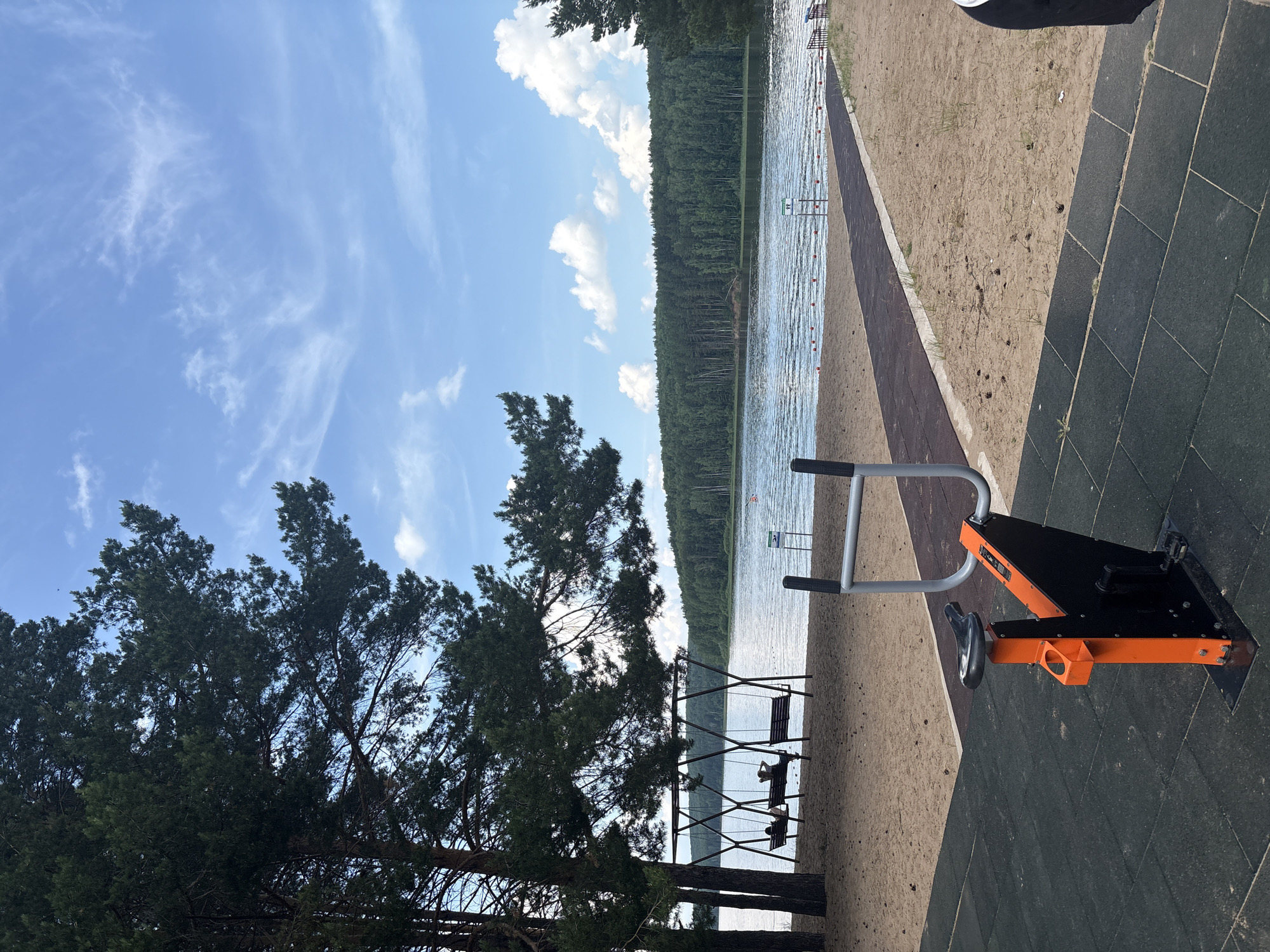 The image shows a public park with a beach and a lake. In the foreground, there is an outdoor exercise machine with an orange and black frame and a black seat, located on a dark rubberized surface. To the left of the exercise machine, a sandy area features three swings, each with two seats. Two of the swings are occupied by individuals sitting facing the lake. Beyond the beach is a calm body of water, the Kama River, with a treeline forming the distant horizon. A dark path runs from the foreground towards the lake, bordered by sand. The sky is blue with scattered white clouds. The location is a beach area along the Kama River in the city of Chaykovsky, Russia.