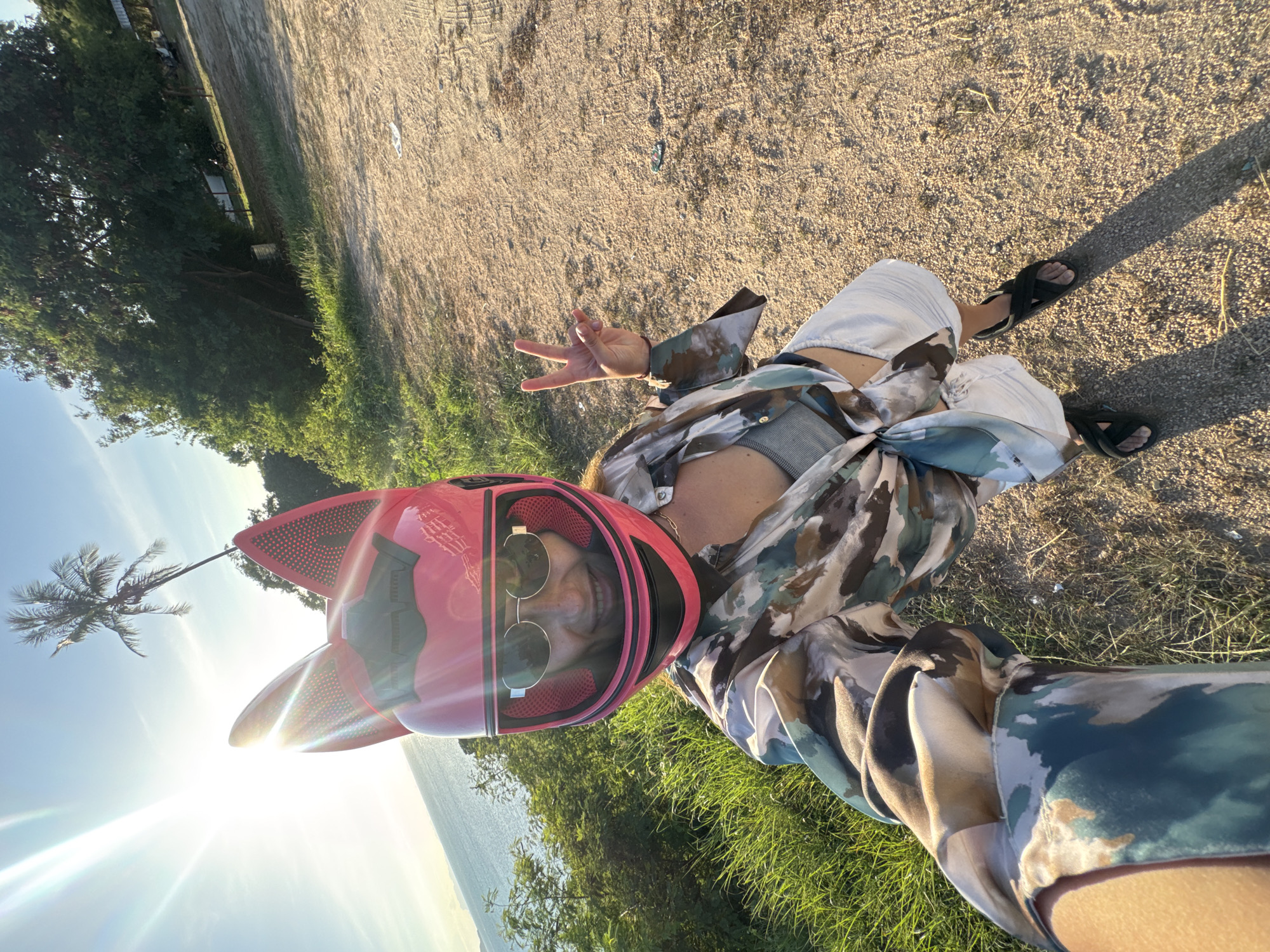 A woman with a pink cat-ear helmet and sunglasses is taking a selfie outdoors. She is wearing a patterned shirt tied at the waist, a gray top, and white shorts. She is also wearing black sandals. She is holding up a peace sign with her right hand. The background includes a body of water, green grass, trees, and a dirt path. The sun is shining brightly.