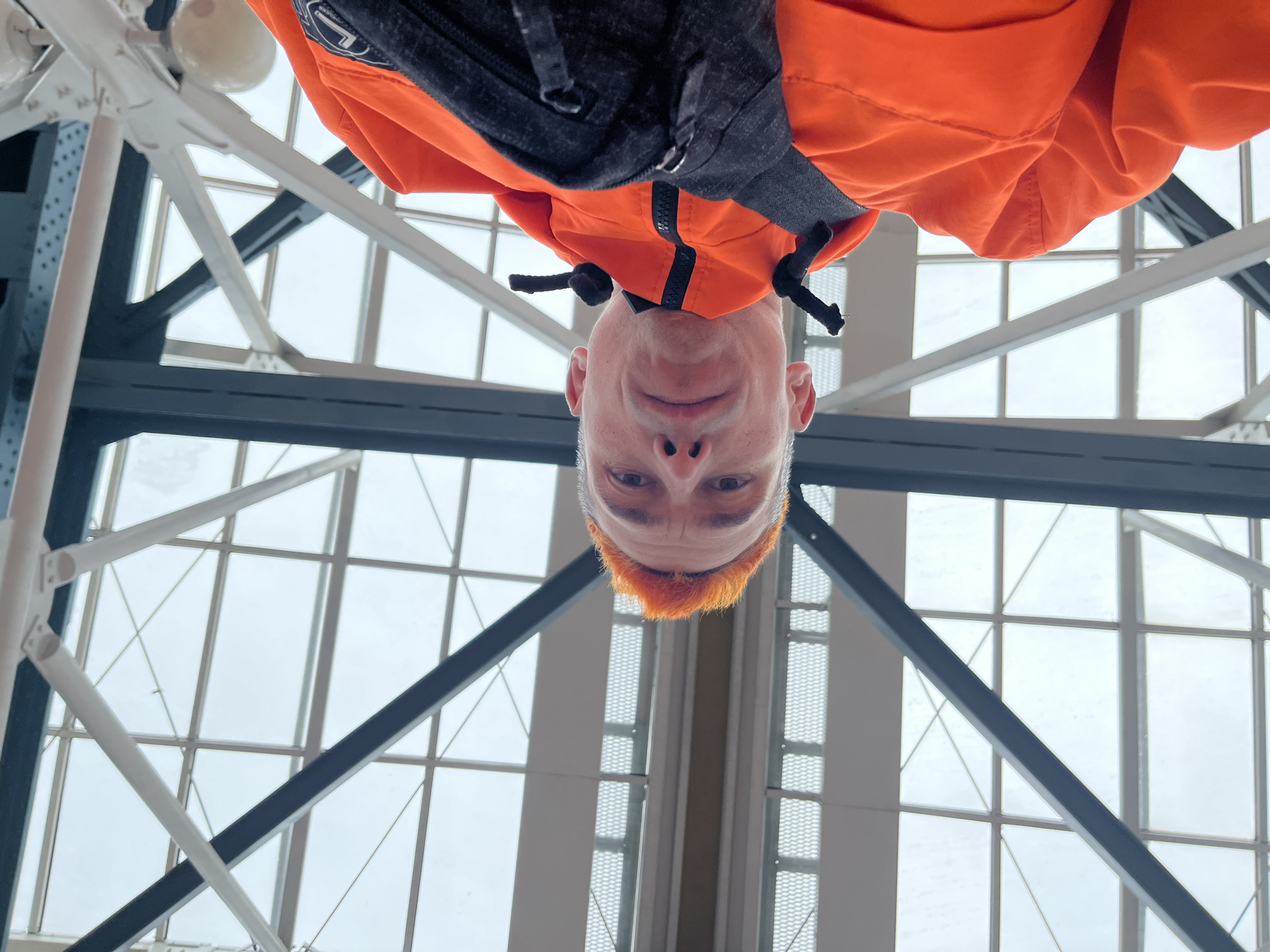 A man with short orange hair is looking down at the camera. He is wearing an orange jacket with a black zippered top underneath. He is standing in a building with a steel grid roof and large windows. The building is located in Moscow, Russia.
