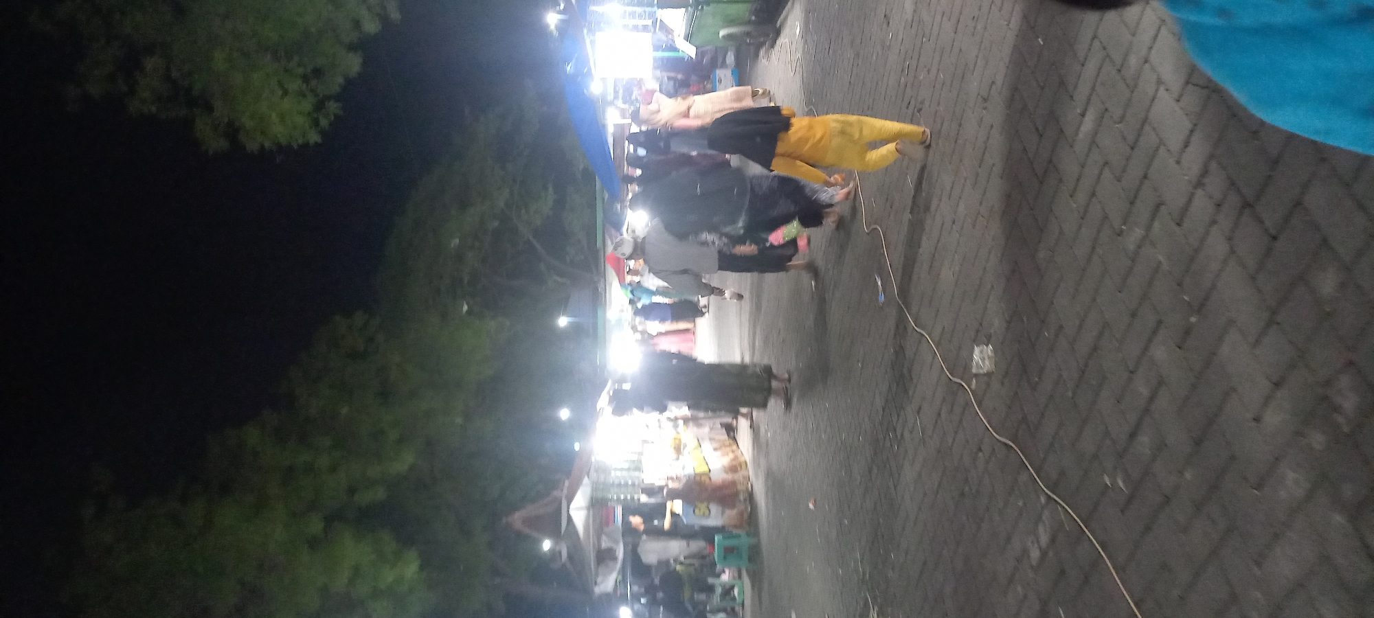 This is a nighttime outdoor photograph taken in the city of Segoro Puro, Indonesia. The scene depicts a market or street fair setting with several stalls and a crowd of people. The ground is paved with dark rectangular bricks, and a light-colored electrical cord runs across it.
In the foreground, a young child wearing a yellow outfit and a black hijab is walking away from the camera. Behind the child, several adults are walking in the same direction. Some are wearing traditional Islamic clothing, such as long robes and head coverings. The people are blurred due to motion or the quality of the photograph.
Further back, there are market stalls illuminated by bright lights, suggesting the sale of goods. Some stalls have awnings or canopies. Trees with green foliage are visible in the background, bordering the area and silhouetted against the dark night sky. Several streetlights are also visible, casting a glow on the scene. The overall atmosphere appears to be lively, characteristic of a bustling night market.