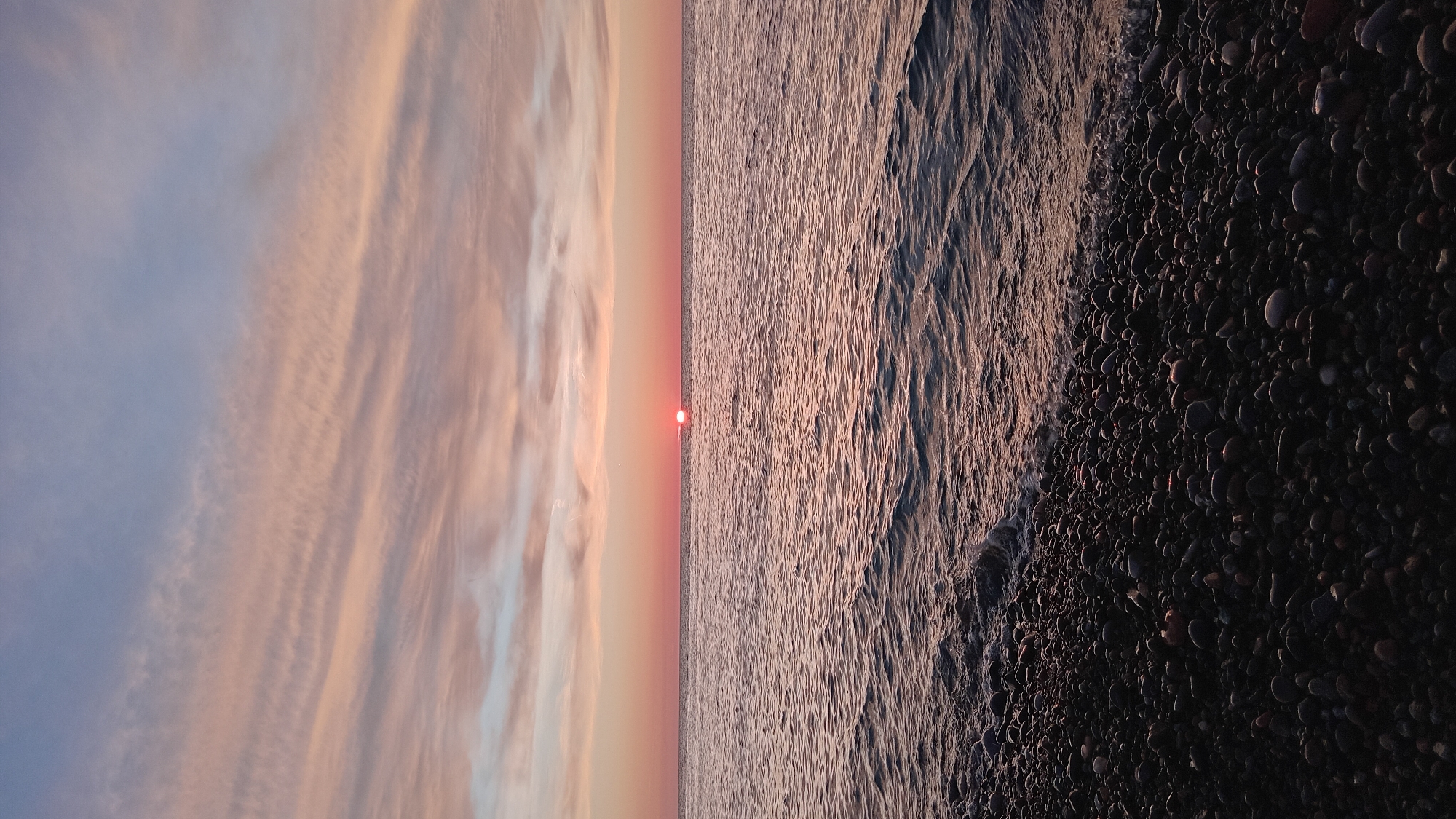 A vertical image depicts a sunset over the Black Sea near Batumi, Georgia. The horizon line bisects the frame horizontally, with a prominent, reddish-orange sun disc visible directly on the horizon. The sky above features horizontal bands of pale pink, orange, and light blue altocumulus clouds. The sea surface is characterized by calm, rippled water reflecting the sky's subdued colors in varying tones of dark blue and muted pink. The foreground consists of a densely packed pebble beach, composed of numerous dark, rounded stones. Small, white-crested waves are observed gently washing onto the pebble shoreline. No individuals, animals, or man-made objects are visible within the scene.