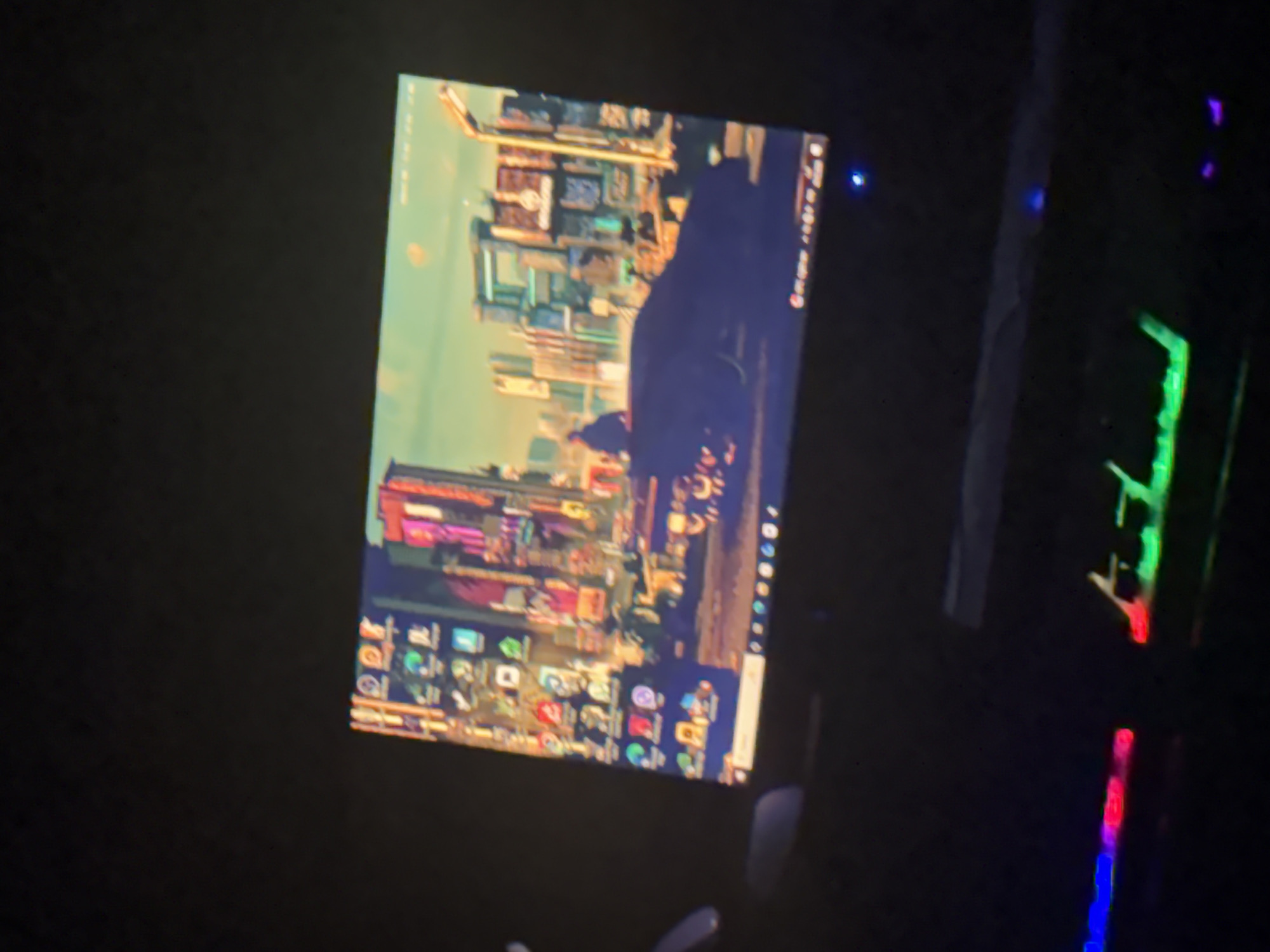 The image shows a computer screen displaying a city background, presumably on the desktop. Icons are visible on the lower part of the screen. To the right of the screen, a keyboard with RGB lighting is partially visible. The environment surrounding the computer setup appears dark. There is no indication that this image is in Yerevan, Armenia.