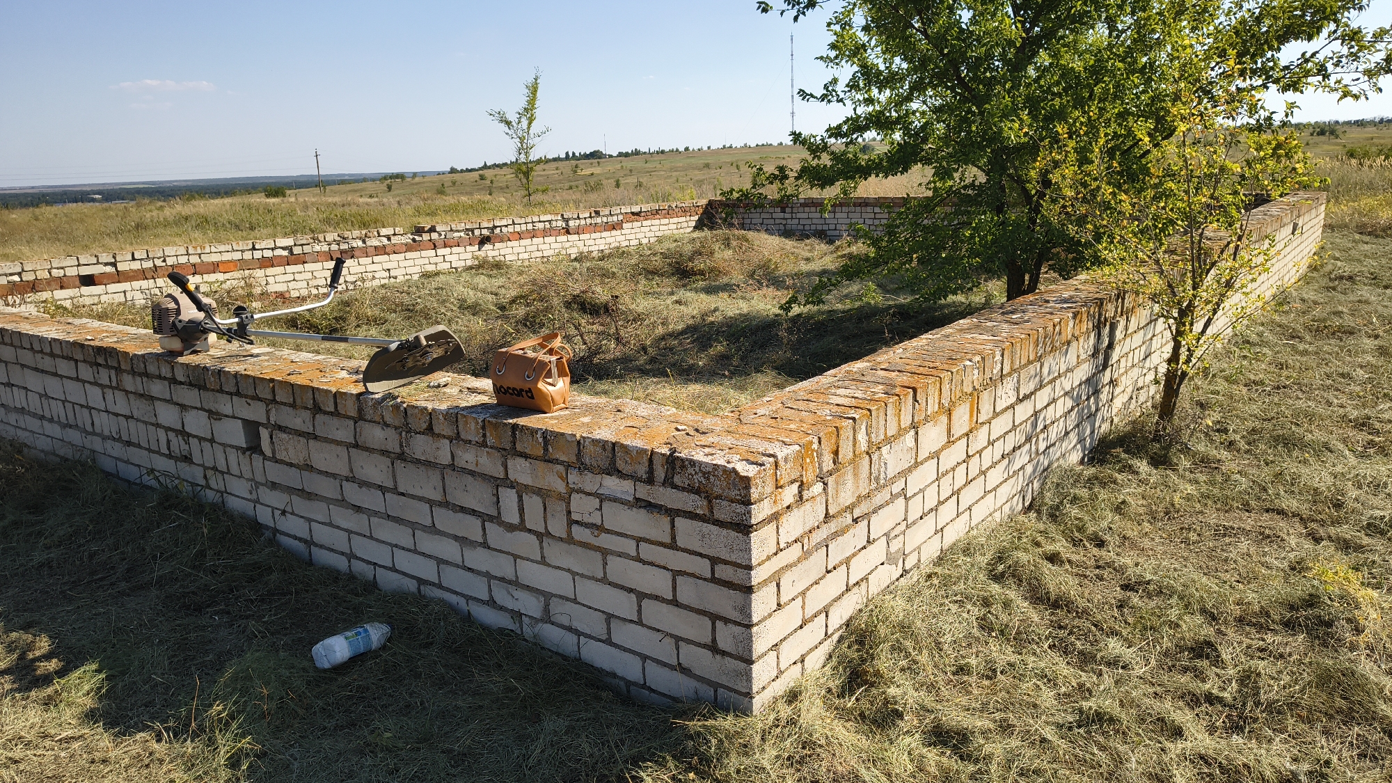An unfinished brick foundation wall, partially overgrown with dry grass and weeds, stands in an outdoor rural landscape. A brush cutter is positioned on top of the wall, along with a small brown bag. A plastic bottle lies on the ground near the wall. The scene is set under a clear blue sky with a tree providing shade on the right side. In the distance, a treeline and rolling hills are visible, along with a distant radio tower. The location is Mikhaylovka, Russia.