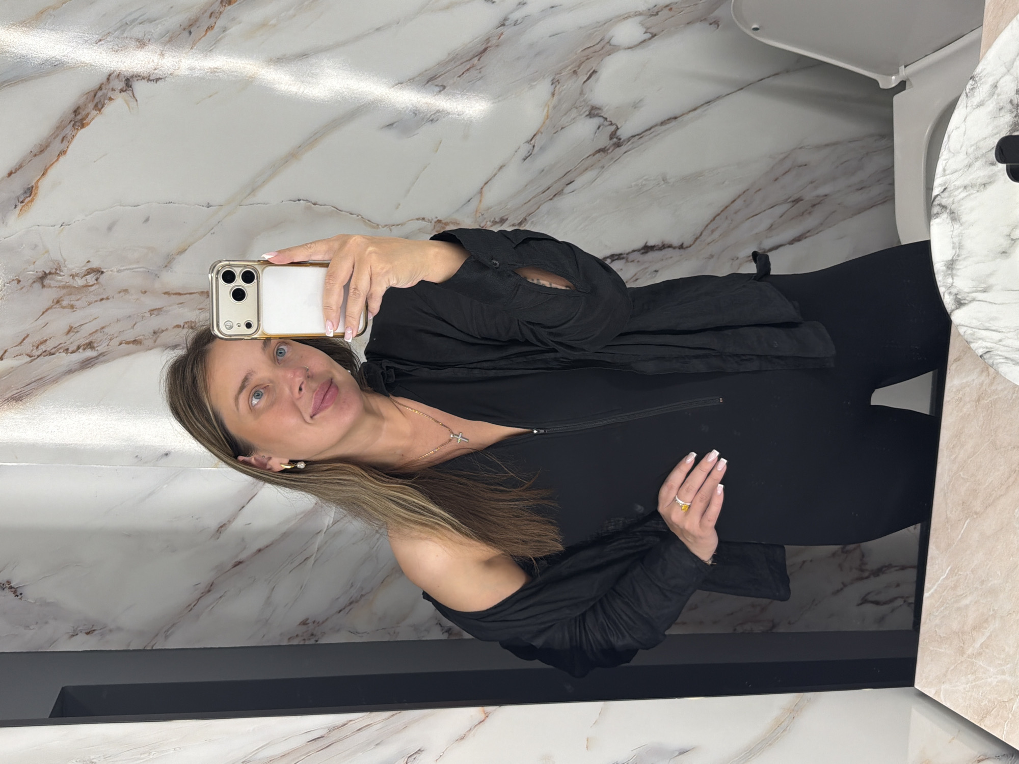 A woman is taking a mirror selfie in a bathroom. She is wearing a black jumpsuit and a black open button-up shirt draped over her shoulders. She has long blonde hair and is wearing gold hoop earrings and a gold necklace with a cross pendant. She is also wearing a gold ring with a large yellow stone on her left hand. The bathroom has marble-patterned walls and a white toilet is visible to the right. The floor in the foreground is a pinkish-white marbled countertop. The image appears to have been taken in the city of Ykremnivs'ke, Ukraine.