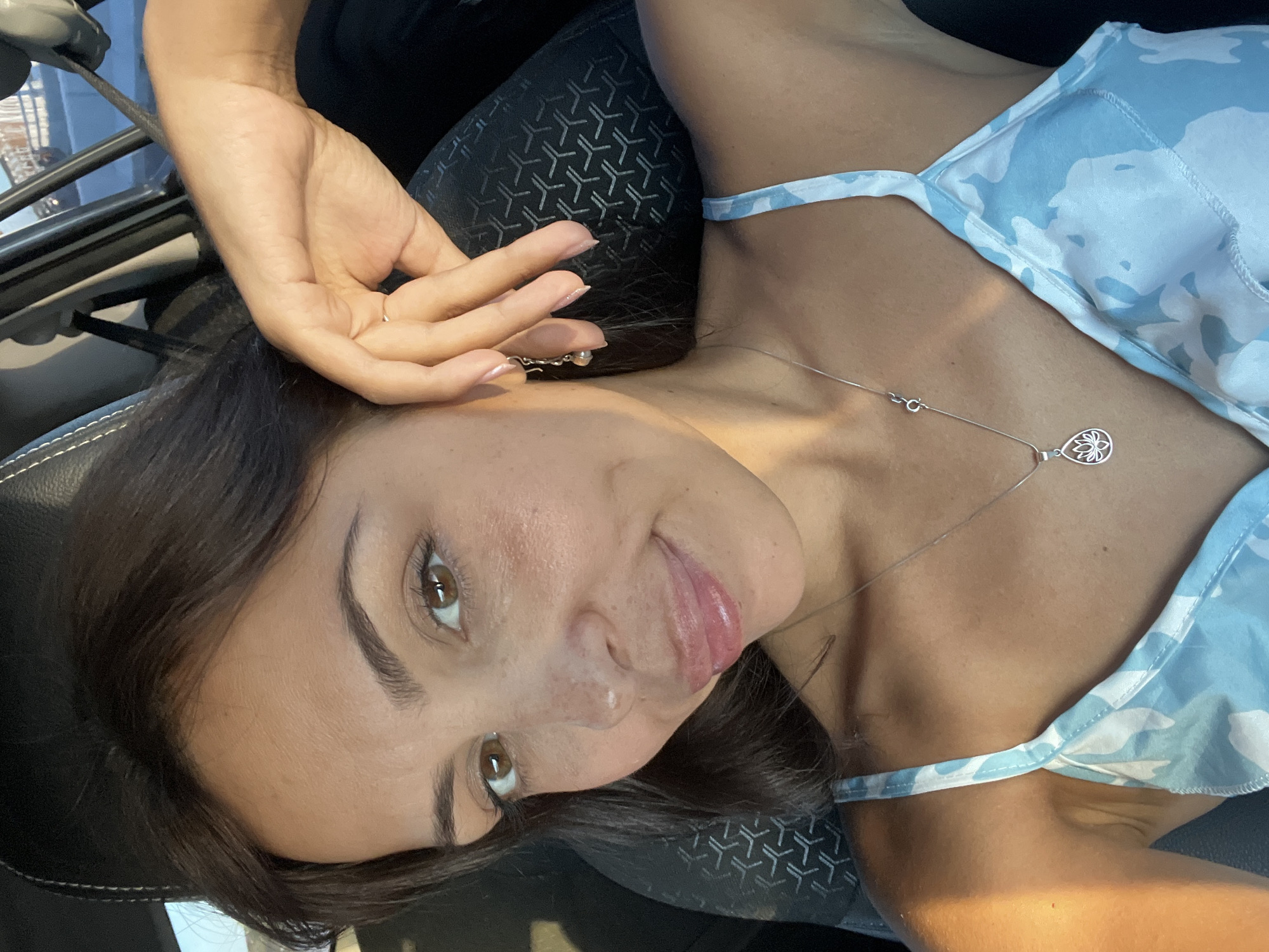 A young woman with brown hair and light brown eyes poses for a selfie. She is indoors, possibly in a car, as evidenced by the car seat texture and the reflection of what appears to be buildings outside. She is wearing a blue and white patterned camisole. She has a silver necklace with a teardrop-shaped pendant. She has one hand raised near her face. The location is not definitively verifiable as Kuta Selatan, Indonesia based on the image alone.