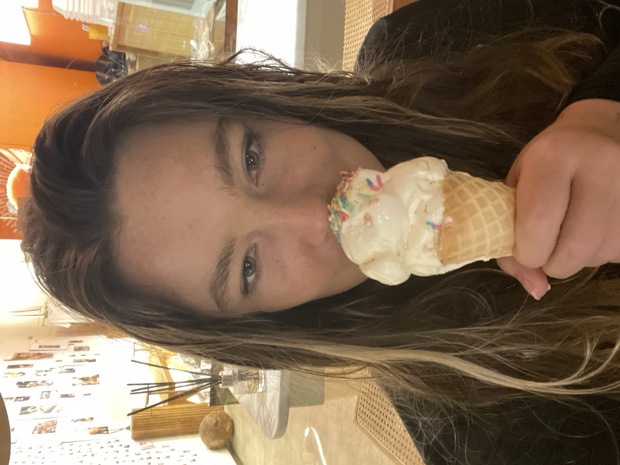 In North Kuta, Indonesia, a young woman with long, dark blonde hair, parted in the center and falling over her shoulders, is pictured in a close-up shot. She is holding a small ice cream cone topped with white ice cream and colorful sprinkles up to her face. Her eyes are looking forward with a neutral expression. The background is out of focus, but appears to be an indoor setting with shelves displaying various items, including photos and decorative objects. The lighting suggests it is daytime or a well-lit interior space.